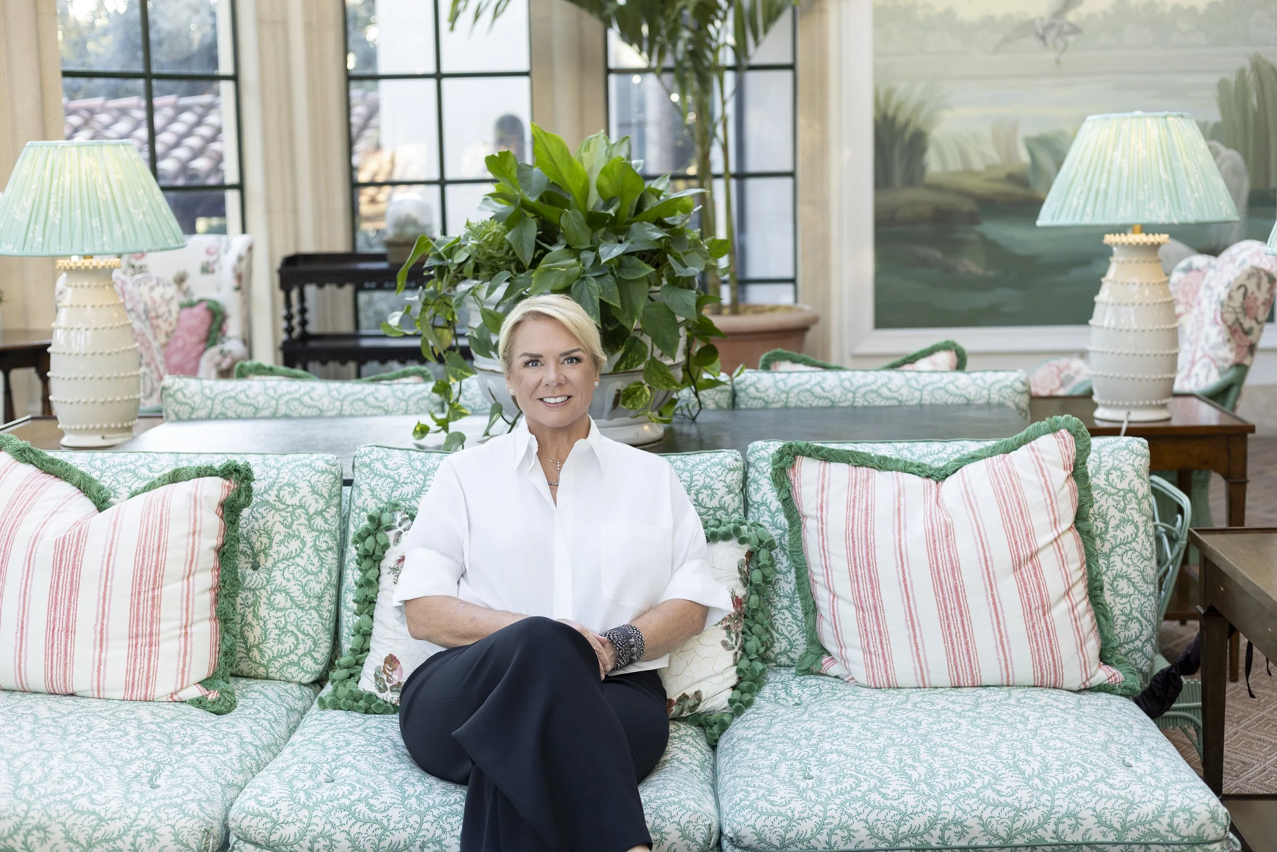 A woman sitting on a patterned sofa in a bright room with large windows, green plants, and lamps on side tables.