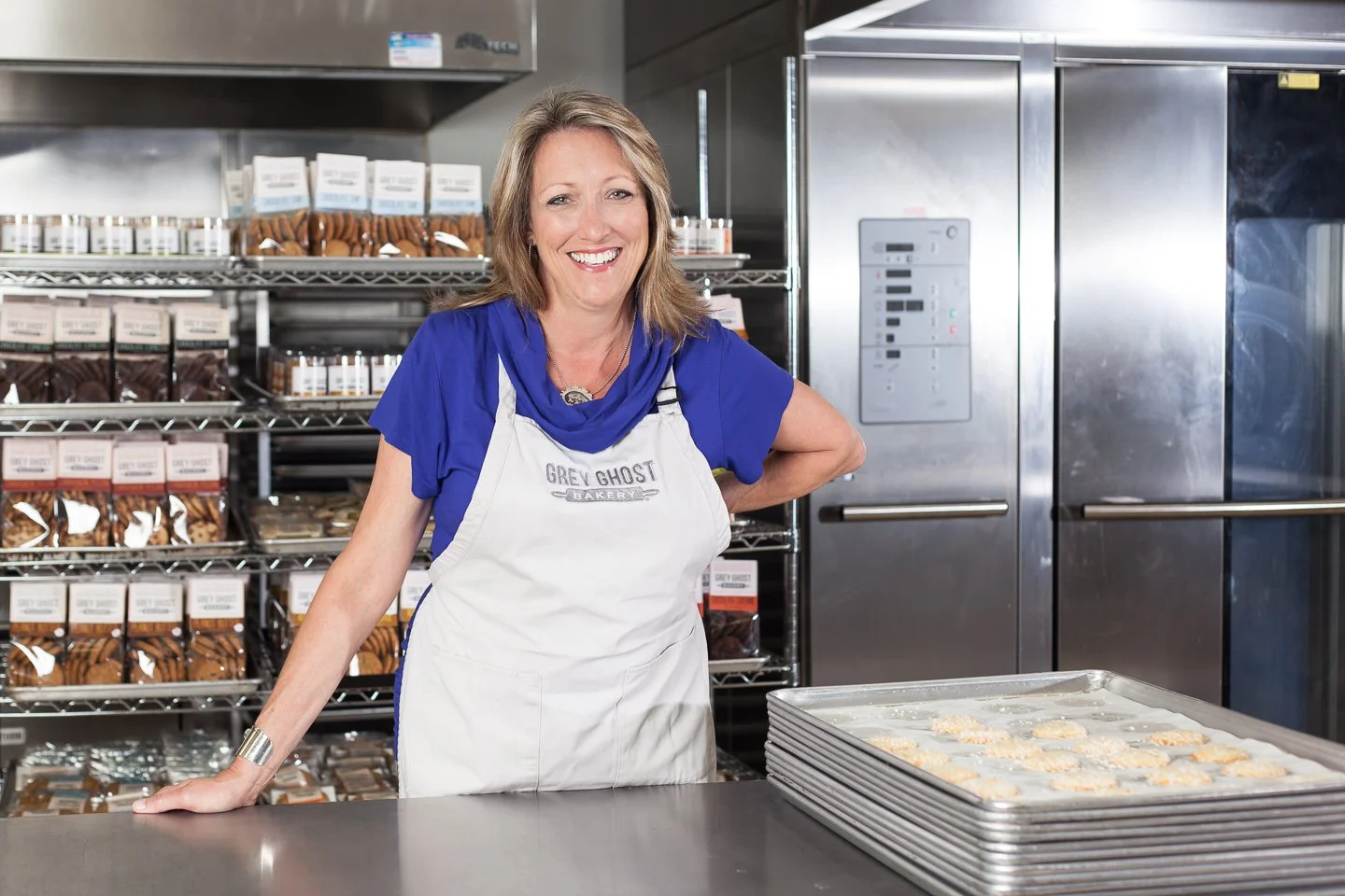 A woman in a white apron with 'Grey Ghost Bakery' logo, standing behind a bakery counter with trays of cookies, in a commercial kitchen with shelves of packaged baked goods and industrial appliances.