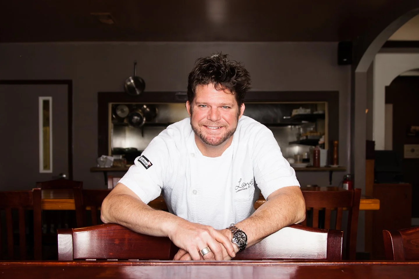 A smiling male chef with dark, curly hair and a beard, wearing a white chef's jacket, leaning on a wooden table in a restaurant kitchen or dining area.