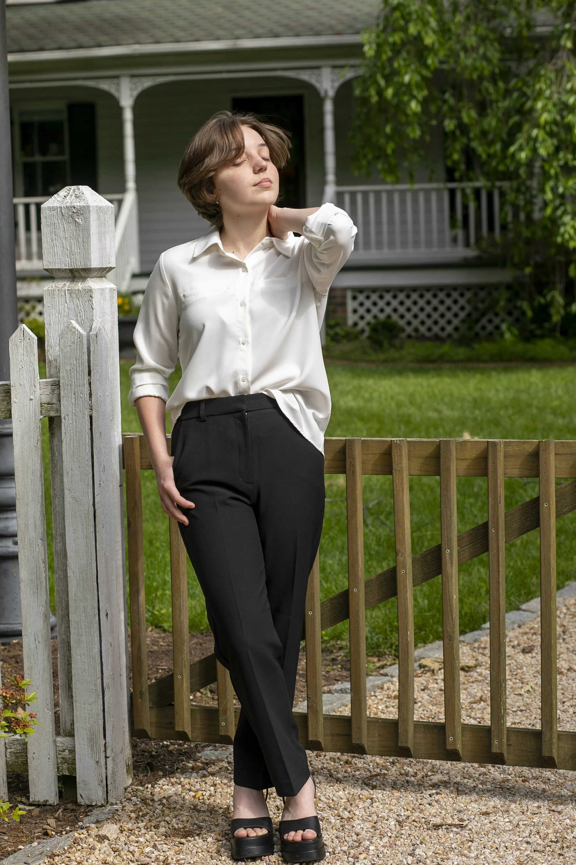A young woman with short brown hair, wearing a white blouse and black pants, standing outdoors near a wooden fence with a house and green lawn in the background.