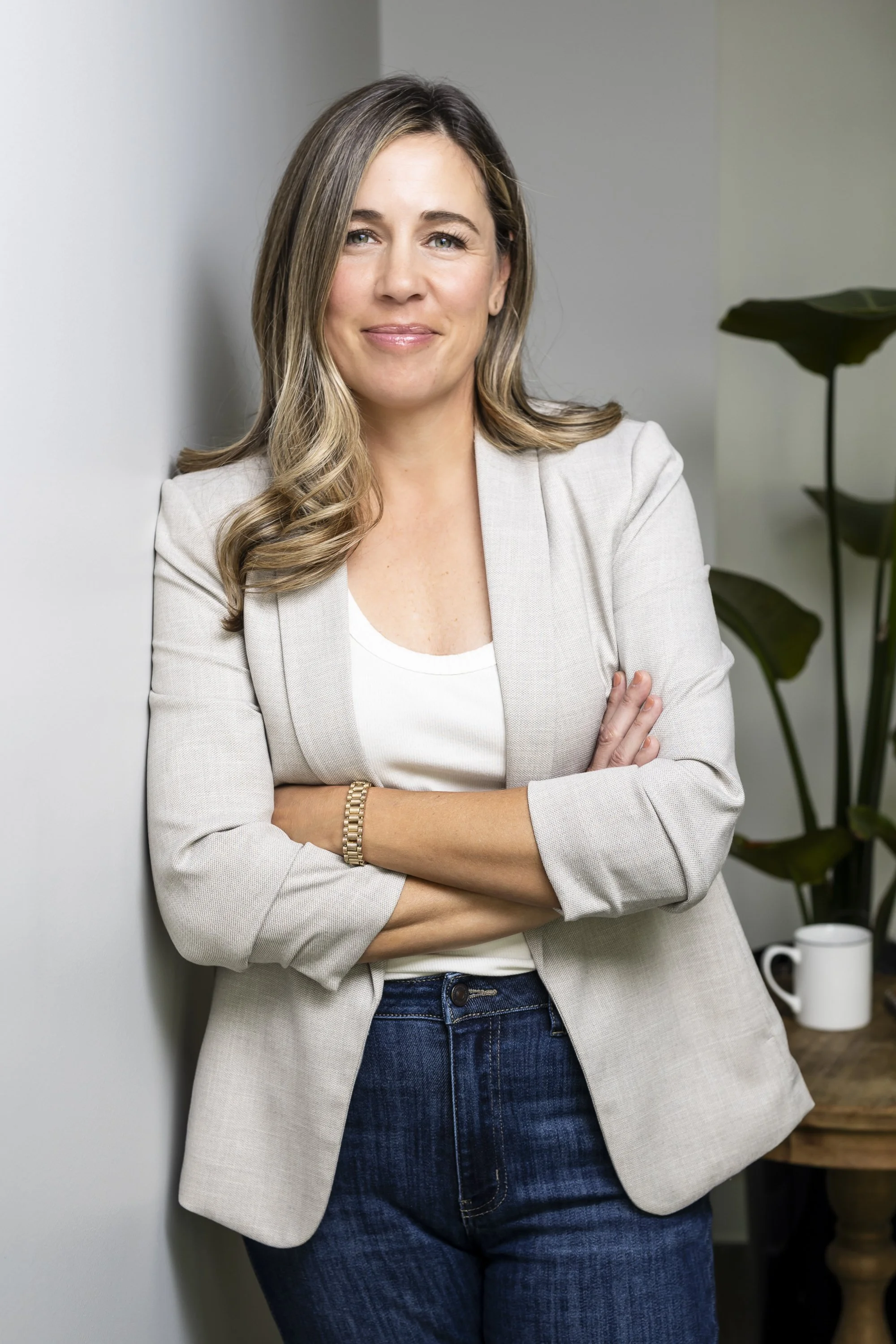 A woman with shoulder-length, wavy hair, wearing a light gray blazer over a white top, standing with arms crossed in an office setting. In the background, there are green plants and a wooden table with a white mug.