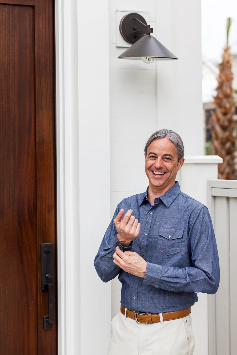 A man with gray hair smiling and gesturing near a front door with a modern wall-mounted lamp.