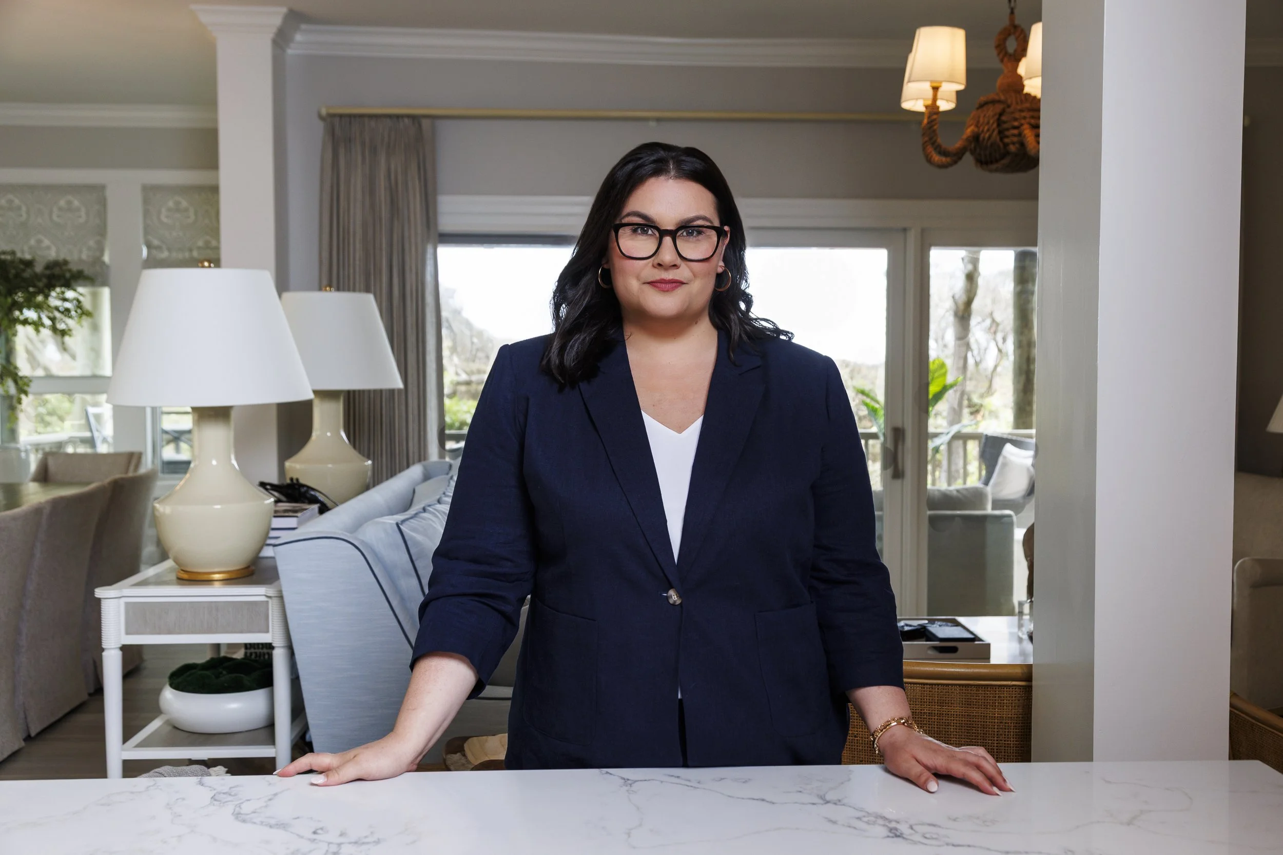 A woman with dark hair, glasses, and a navy blazer standing behind a white marble countertop in a well-lit living room, with window views and modern decorations.