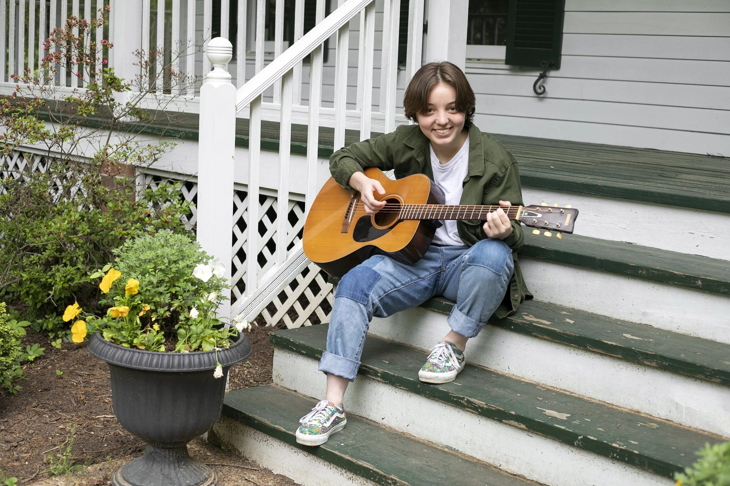 A young person with short brown hair sitting on the steps of a porch, playing an acoustic guitar and smiling, wearing a green jacket, white T-shirt, rolled-up jeans, and colorful sneakers.