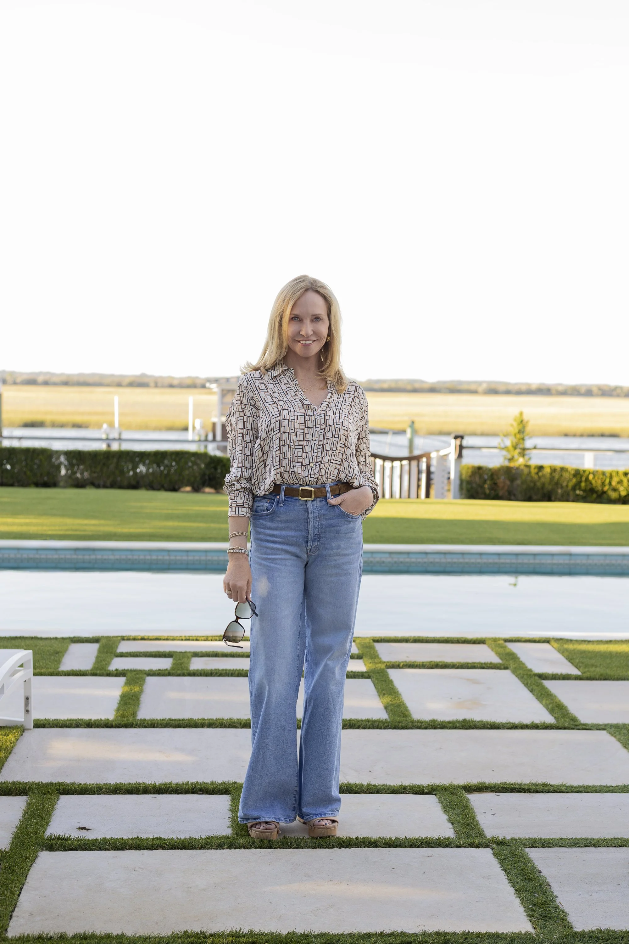 A woman standing on a stone and grass patio near a pool, holding sunglasses, with a lake and open field in the background.