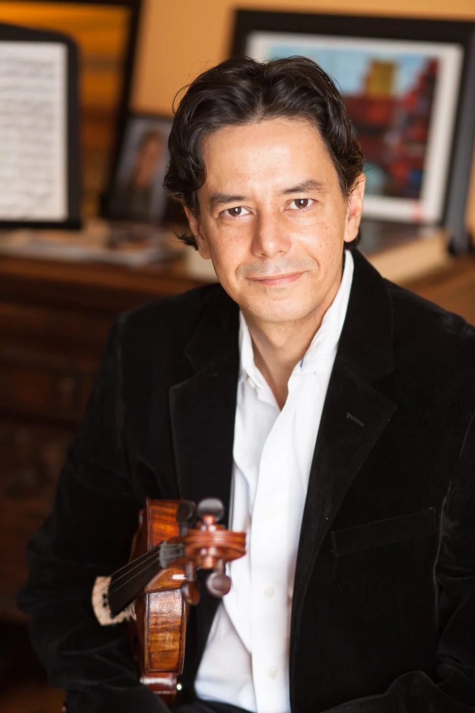A man in a black blazer and white shirt holding a violin, sitting in a room with framed pictures and desktop screens in the background.