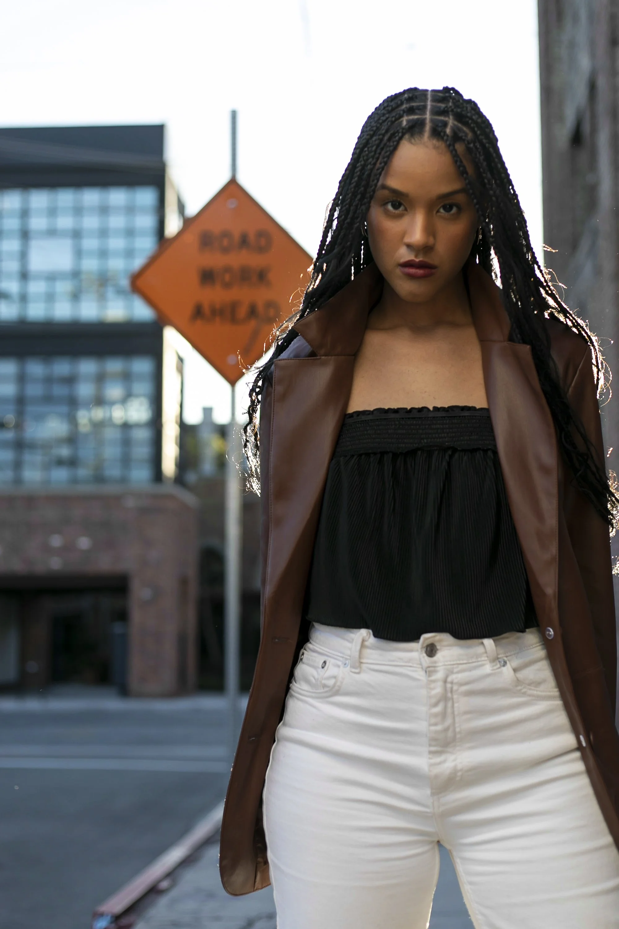 A young woman with long braids standing outdoors in an urban area with a "Road Work Ahead" sign behind her, wearing a brown trench coat, black top, and white pants.