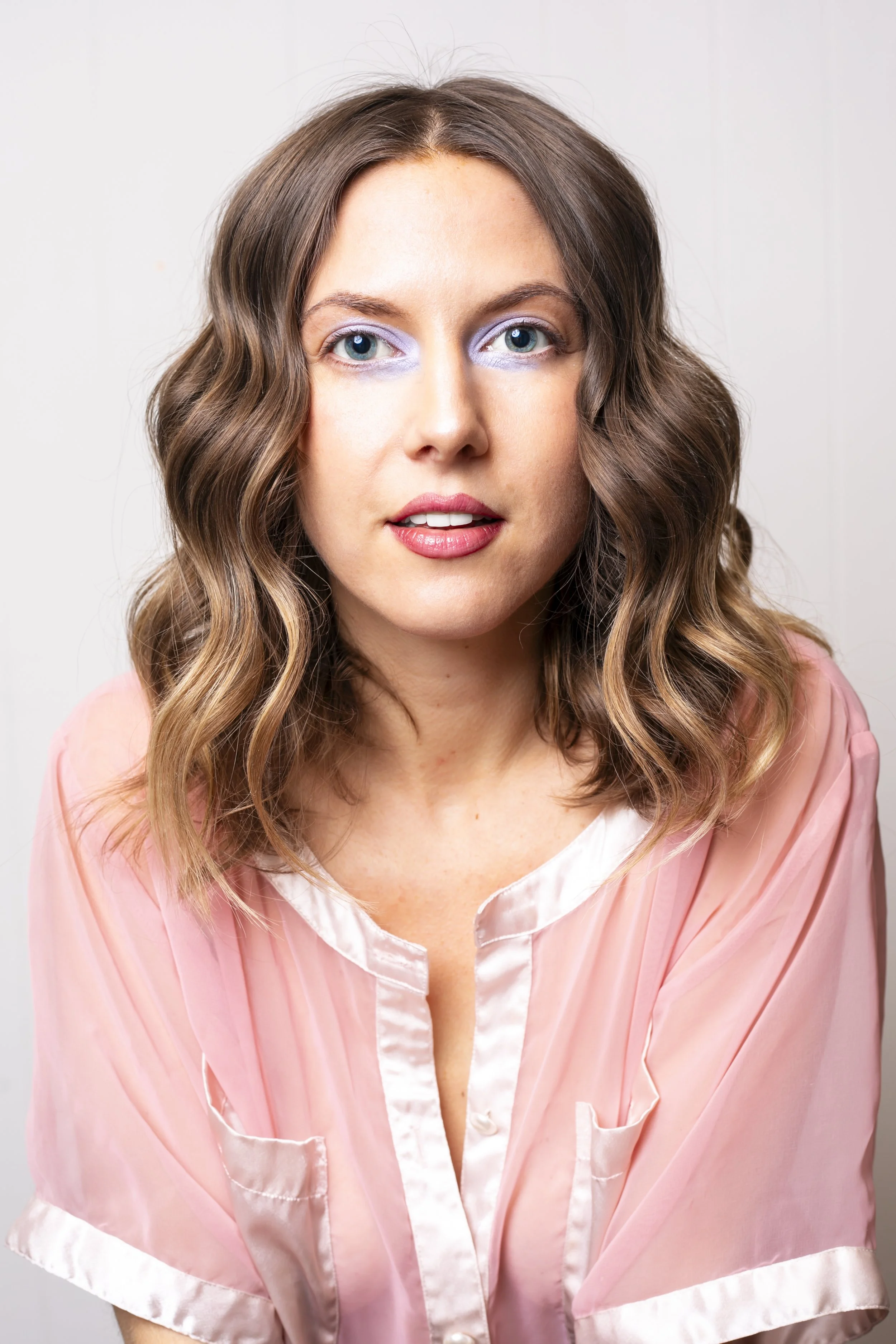 A woman with wavy brown hair, blue eyes, and pink lipstick, wearing a pink silk robe, looking directly at the camera against a plain background.