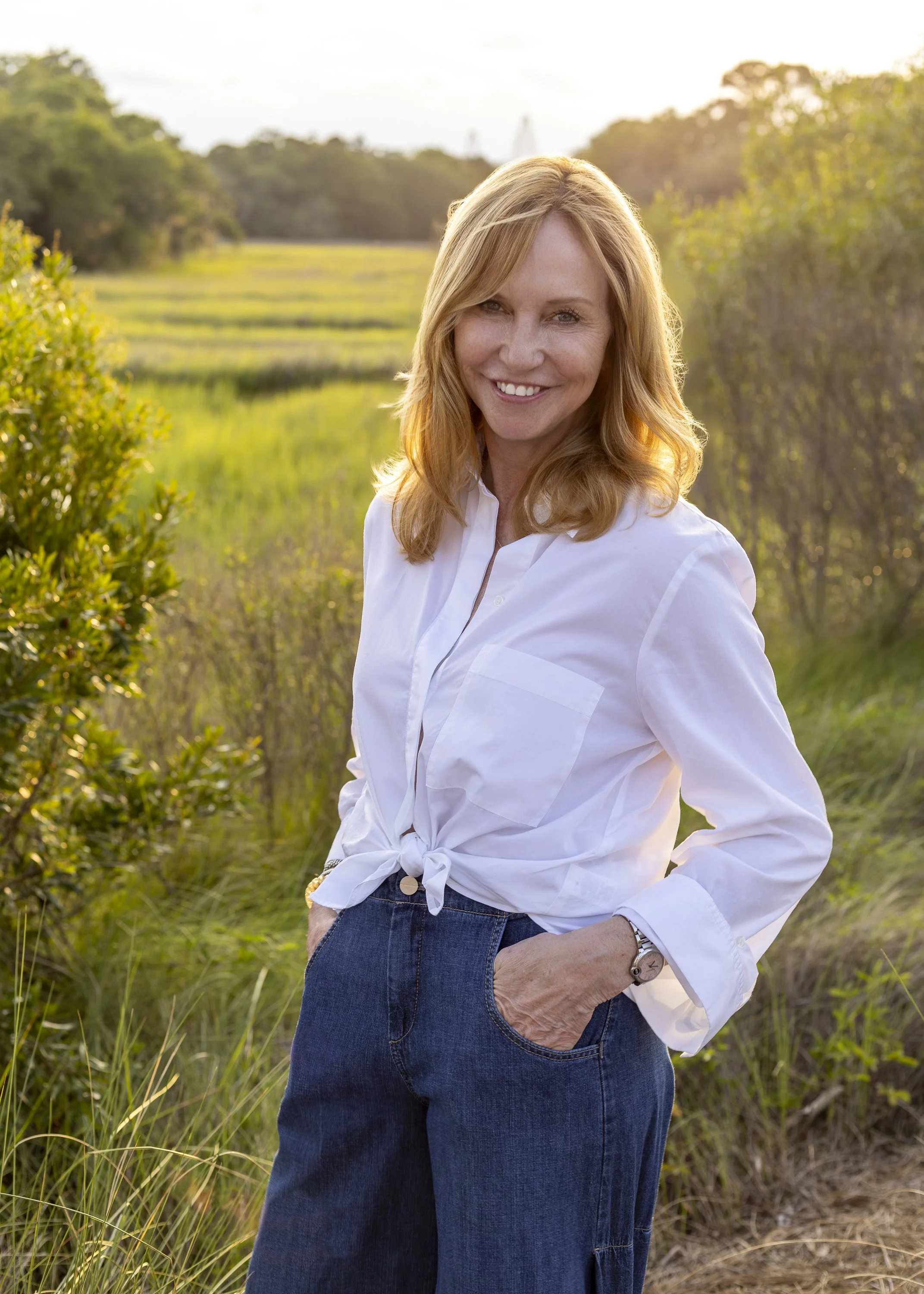 A woman with blonde hair smiling outdoors in a park or nature reserve during sunset, wearing a white shirt tied at the waist and blue jeans.