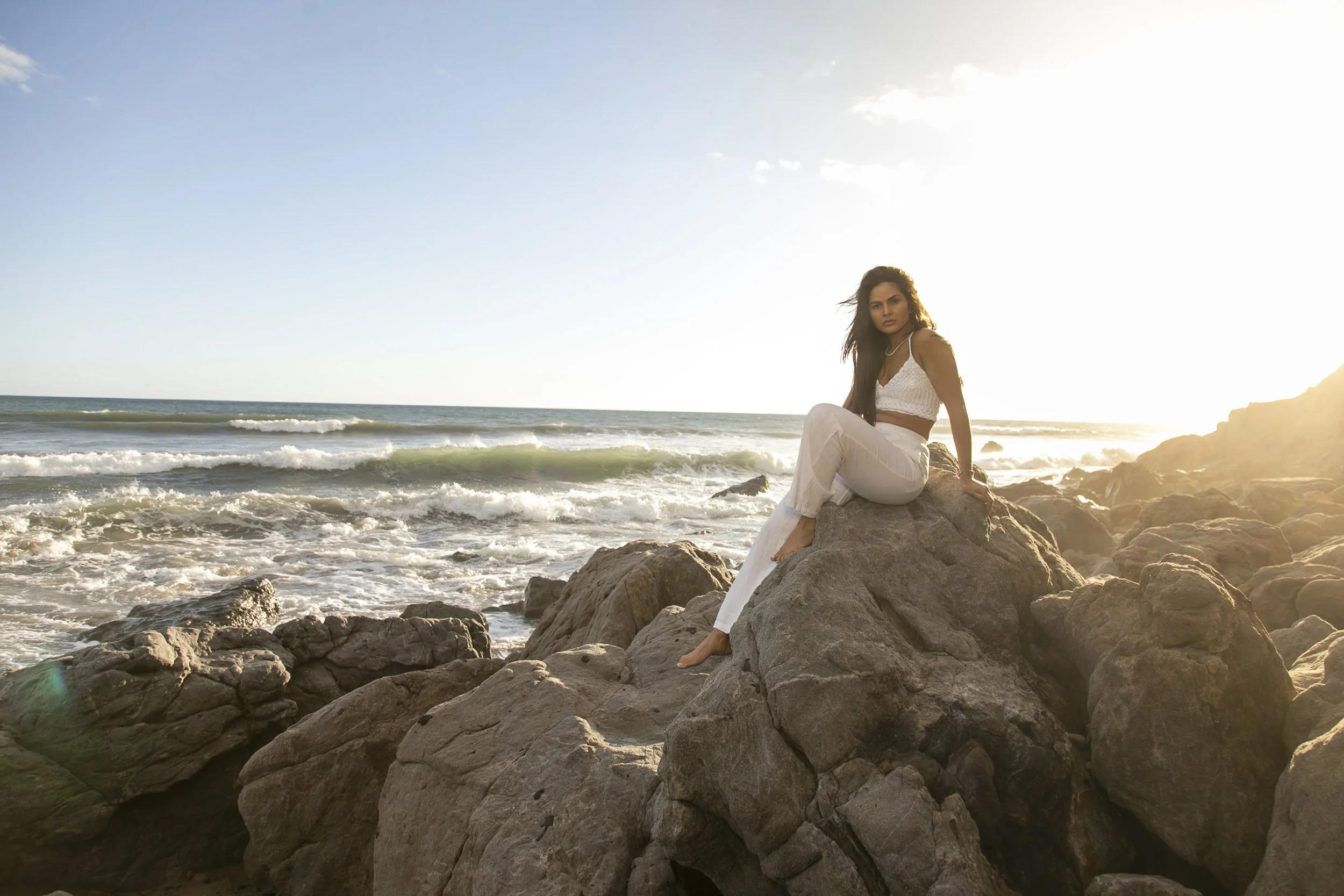 A woman in white clothing sitting on rocks at the beach with ocean waves in the background during sunset.