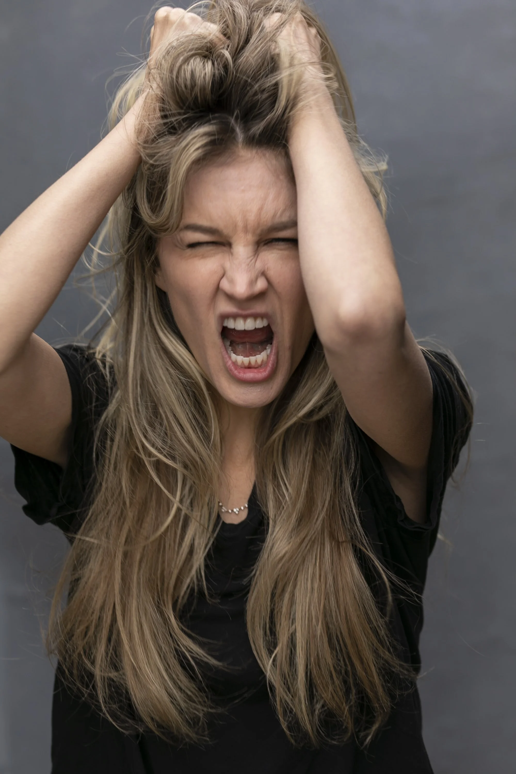 woman with long blonde hair, wearing a black shirt, holding her head with both hands, appearing angry or frustrated