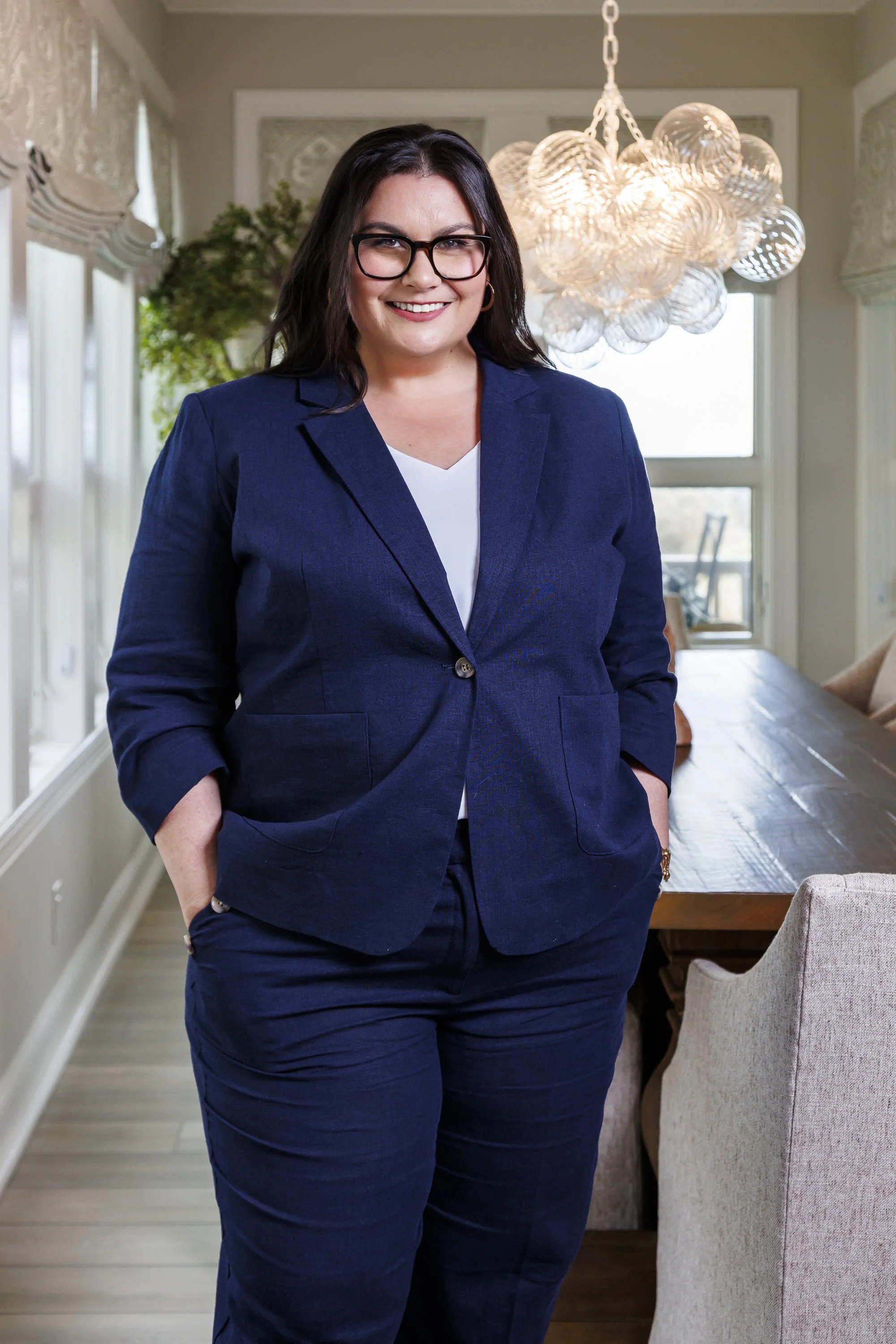 A woman in a blue suit with glasses smiling, standing in a well-lit dining room with a wooden table and modern light fixture.