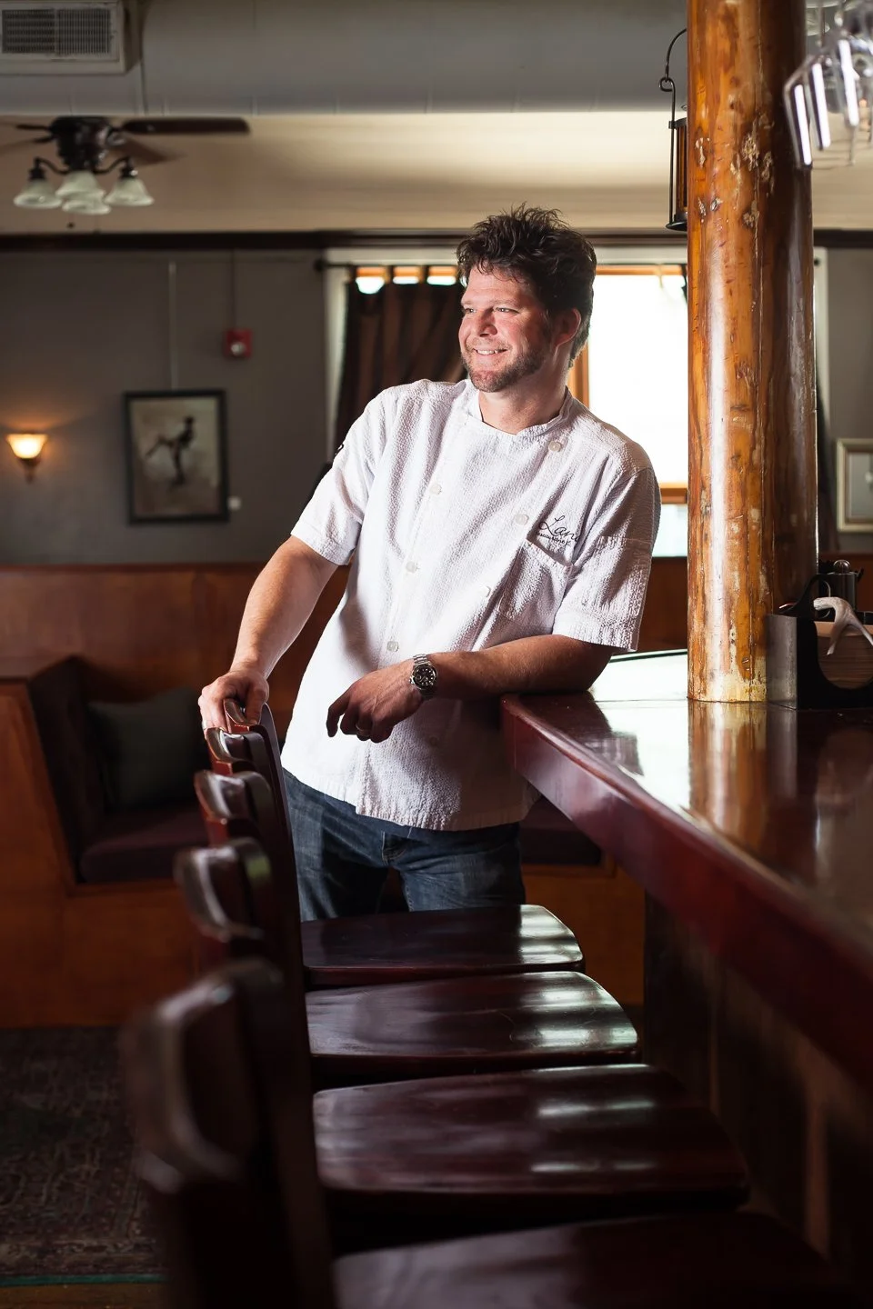 A man in a white chef's coat standing at a bar counter in a restaurant, smiling and looking to the side. The interior has warm wood tones, a window in the background, and a framed picture on the wall.