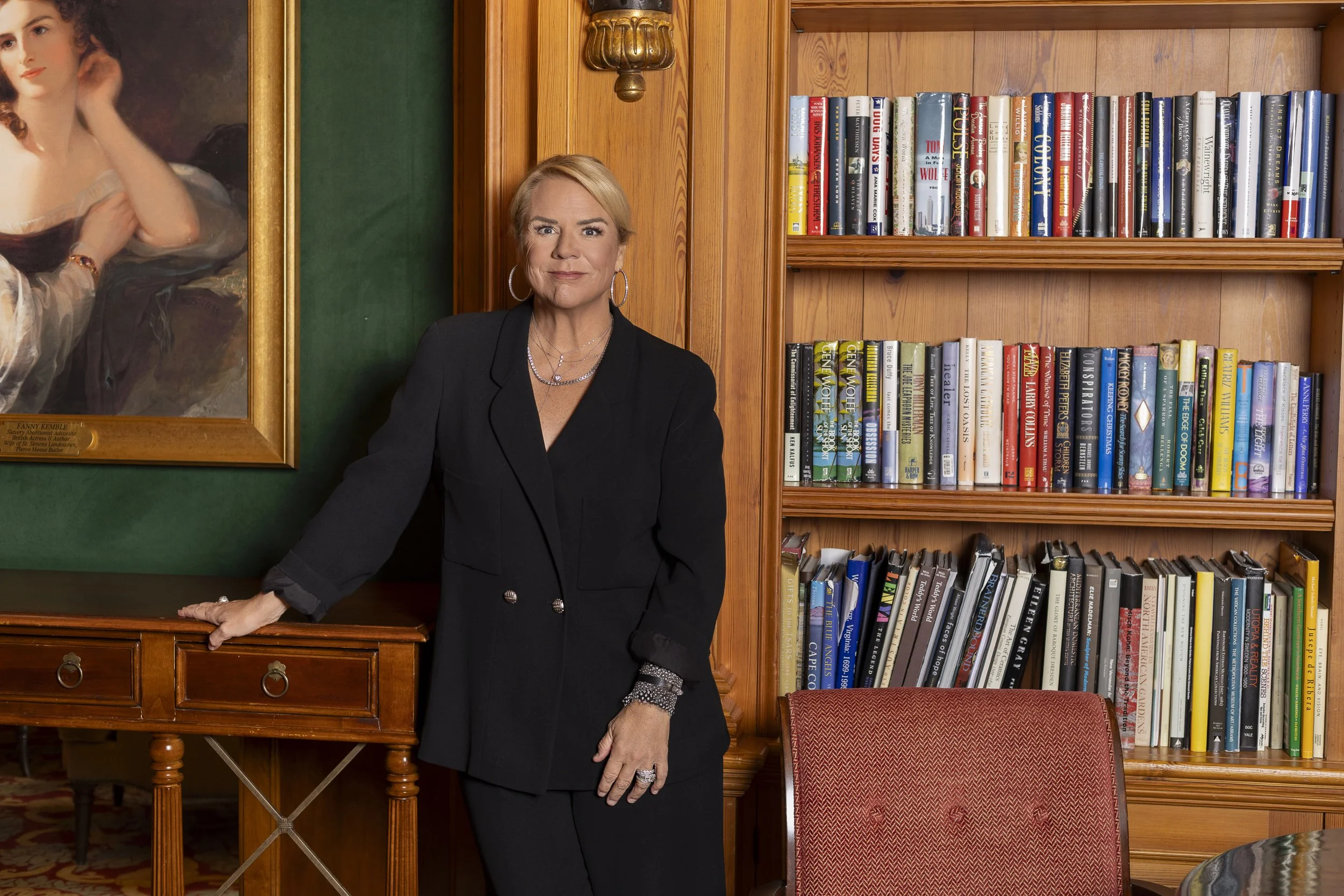 A woman with short blonde hair, dressed in a black blazer and pants, standing in front of a bookshelf filled with books, beside a large portrait of a woman with red hair.