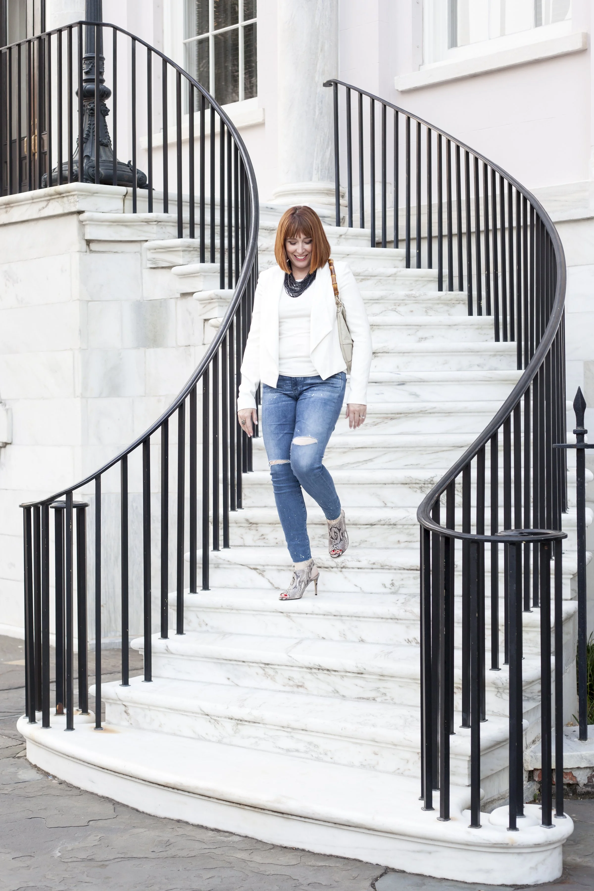 Woman with red hair, white blazer, blue ripped jeans, and snake print high heels walking down curved marble staircase outside a building.