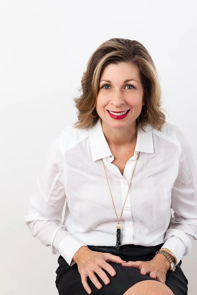 A woman with shoulder-length brown hair, wearing a white blouse, black skirt, gold hoop earrings, and a long necklace with black beads, smiling against a plain white background.