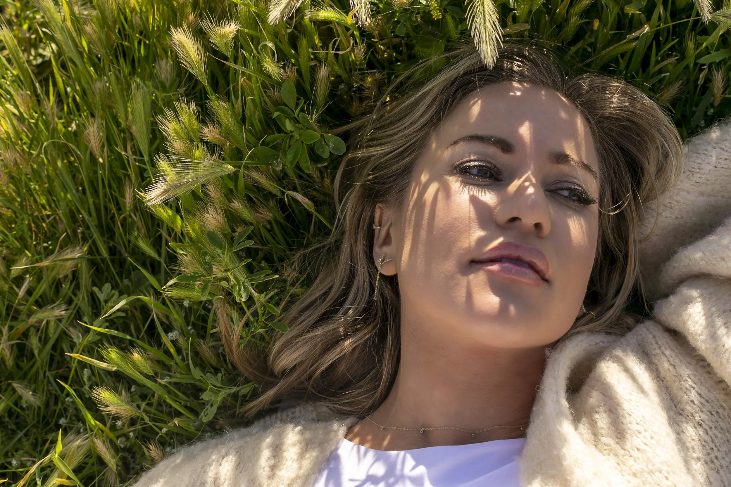 A woman with long light brown hair lies on green grass surrounded by plants and wheat, with shadows of leaves on her face, wearing earrings, a necklace, a white shirt, and a cream-colored sweater.