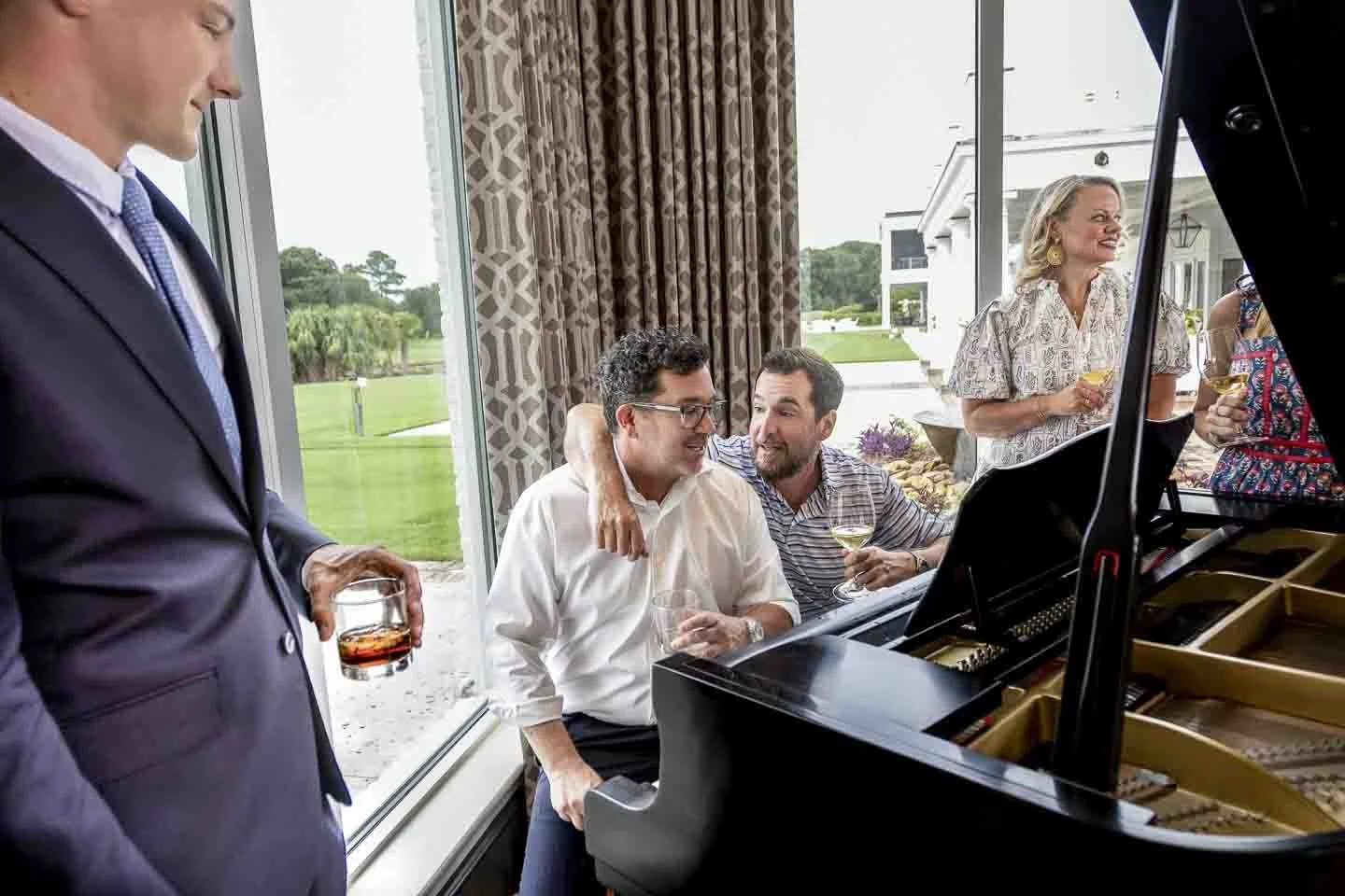 People at a social gathering with a grand piano, some holding drinks, inside a bright room with large windows and a view of a golf course outside.