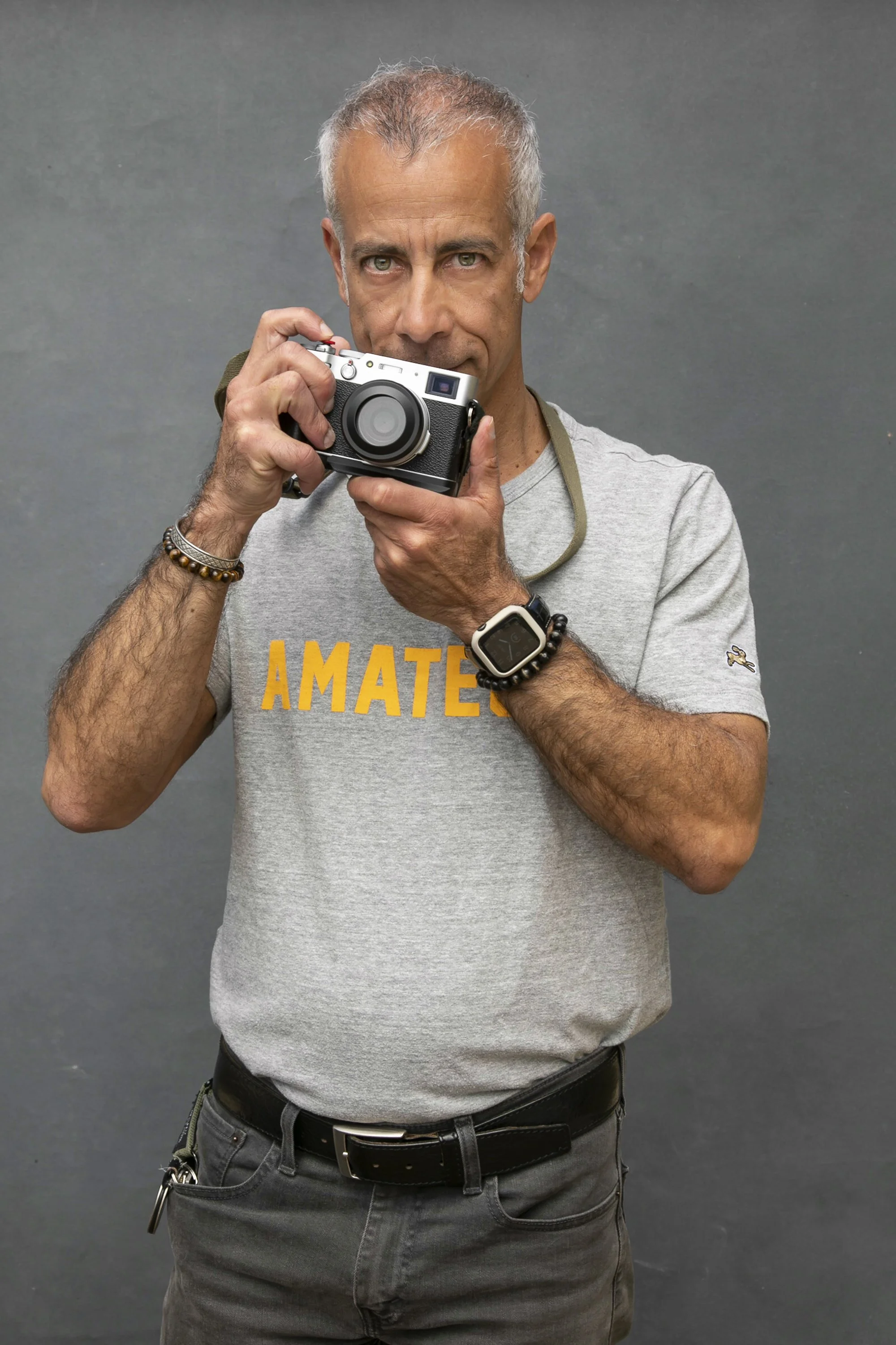 A man with gray hair and blue eyes taking a photograph with a camera, wearing a gray t-shirt that says 'AMATE' and a smartwatch on his wrist, standing in front of a plain gray background.