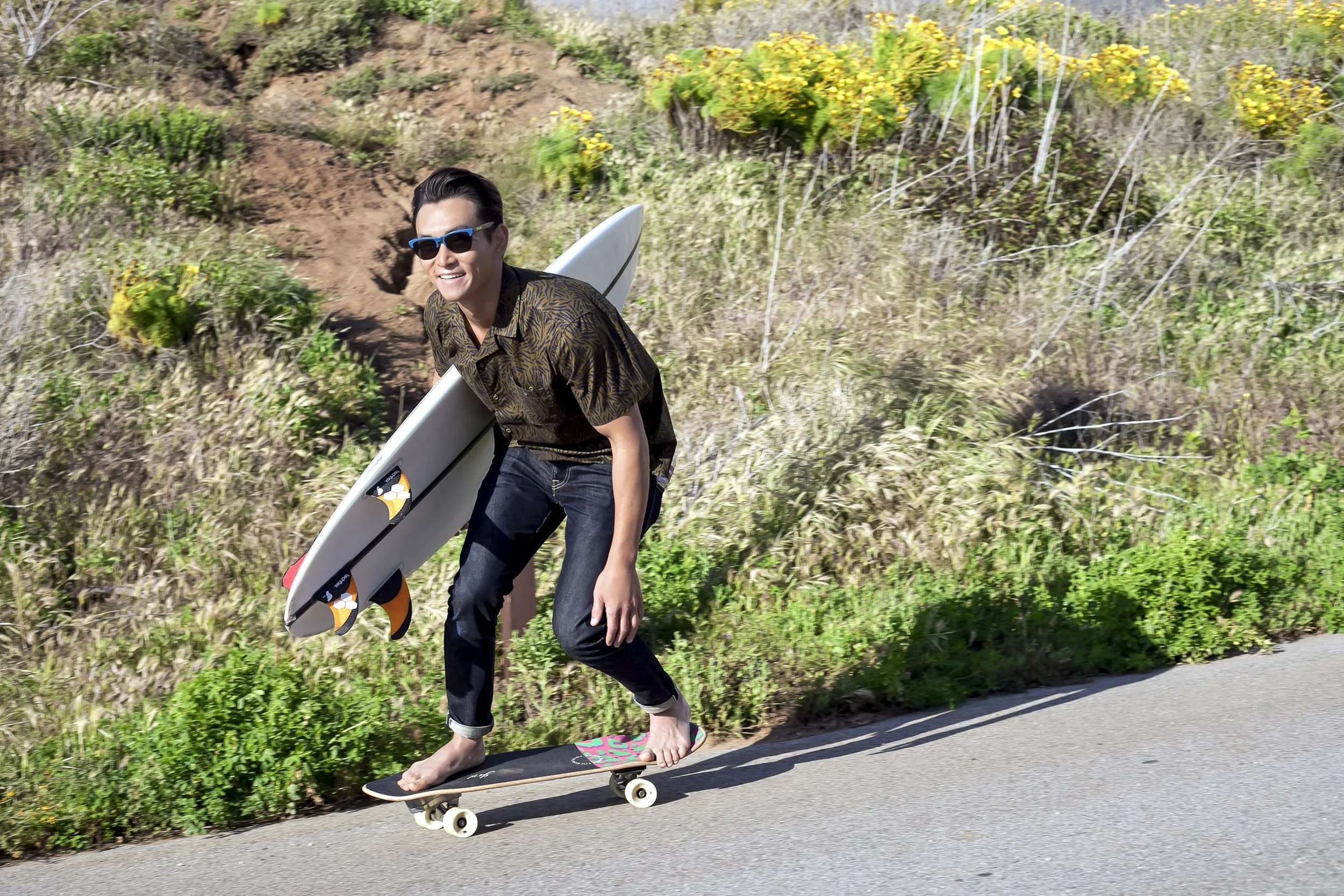 A man with dark hair, wearing sunglasses, a patterned brown shirt, and rolled-up jeans, rides a skateboard while carrying a surfboard on his back along a paved road with grassy and shrub-filled terrain in the background.