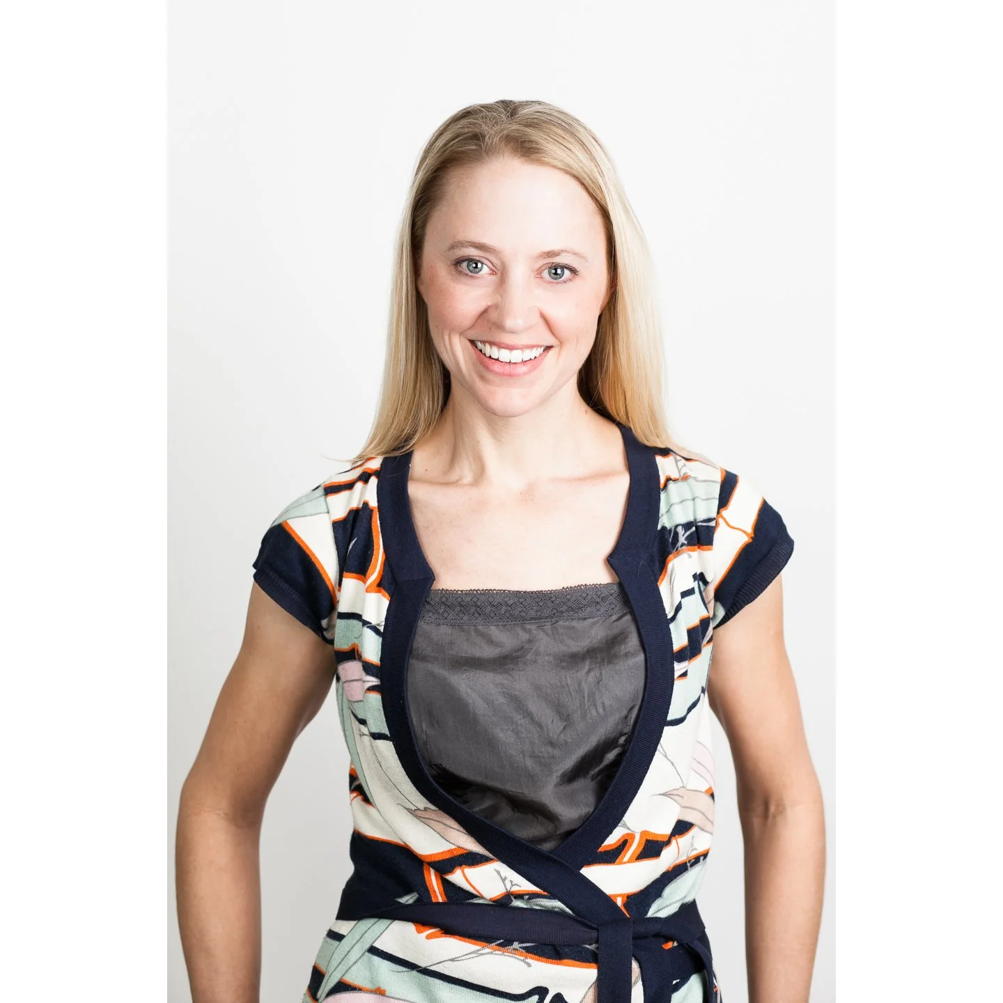 A woman with long blonde hair smiling, wearing a colorful striped top over a gray shirt, standing against a plain white background.