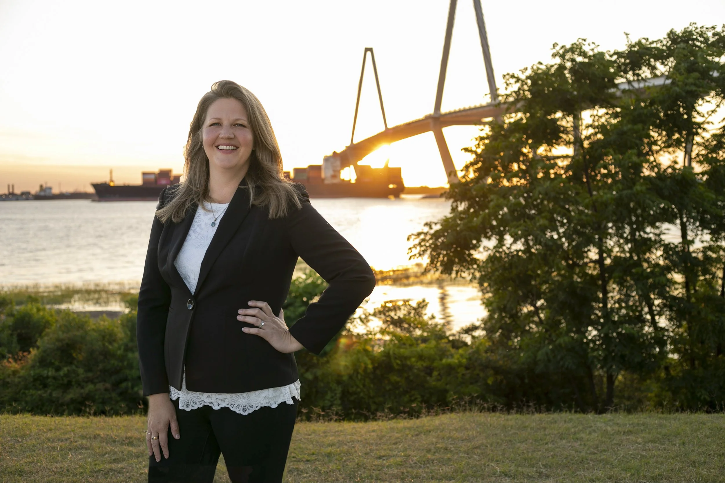 A woman in a black blazer and white top standing outdoors near water during sunset, with a large ship and a crane in the background.