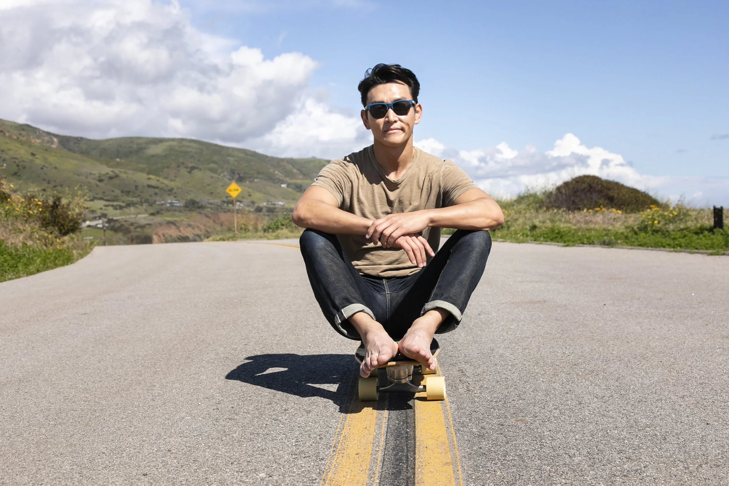 A man sitting cross-legged on a skateboard in the middle of a road with a scenic landscape of hills, clouds, and blue sky in the background.
