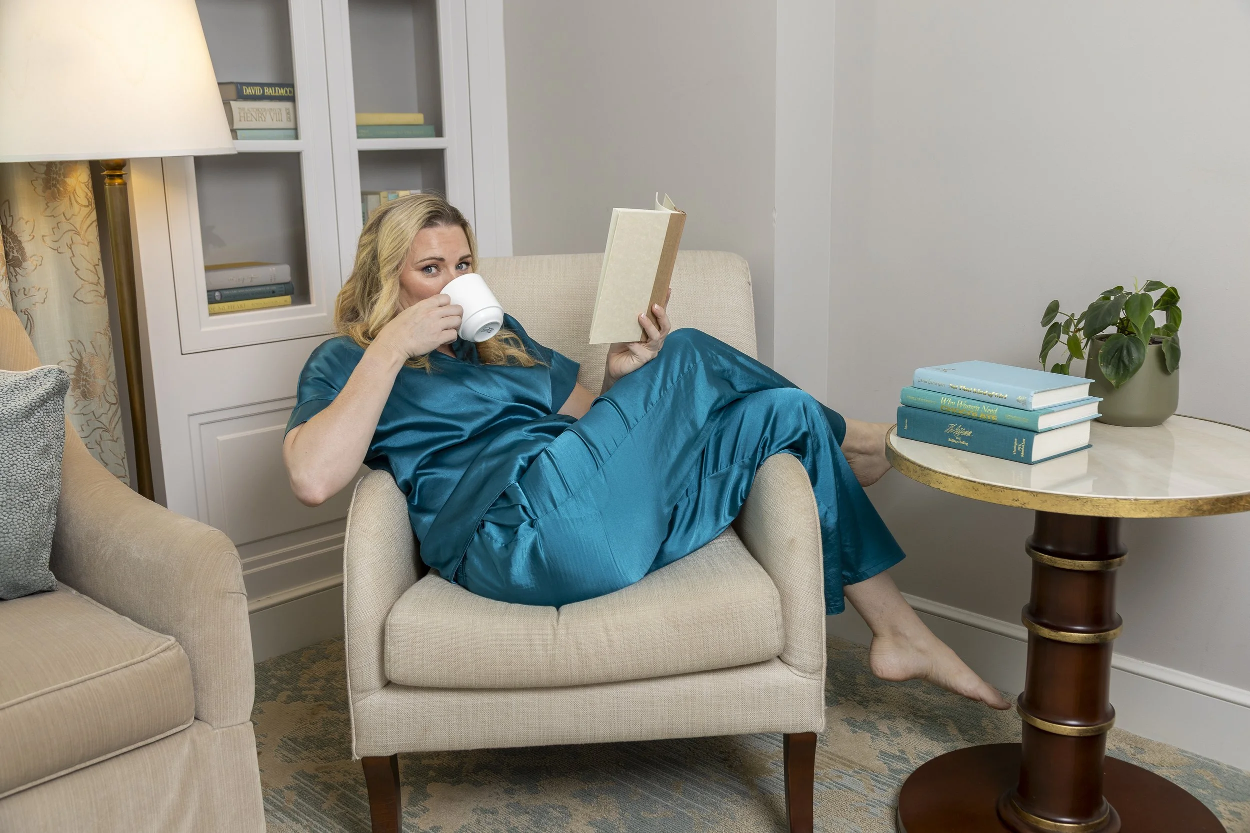 Woman in blue pajamas sitting in an armchair, drinking from a mug and holding a book, in a cozy living room with a side table, plants, and bookshelves.