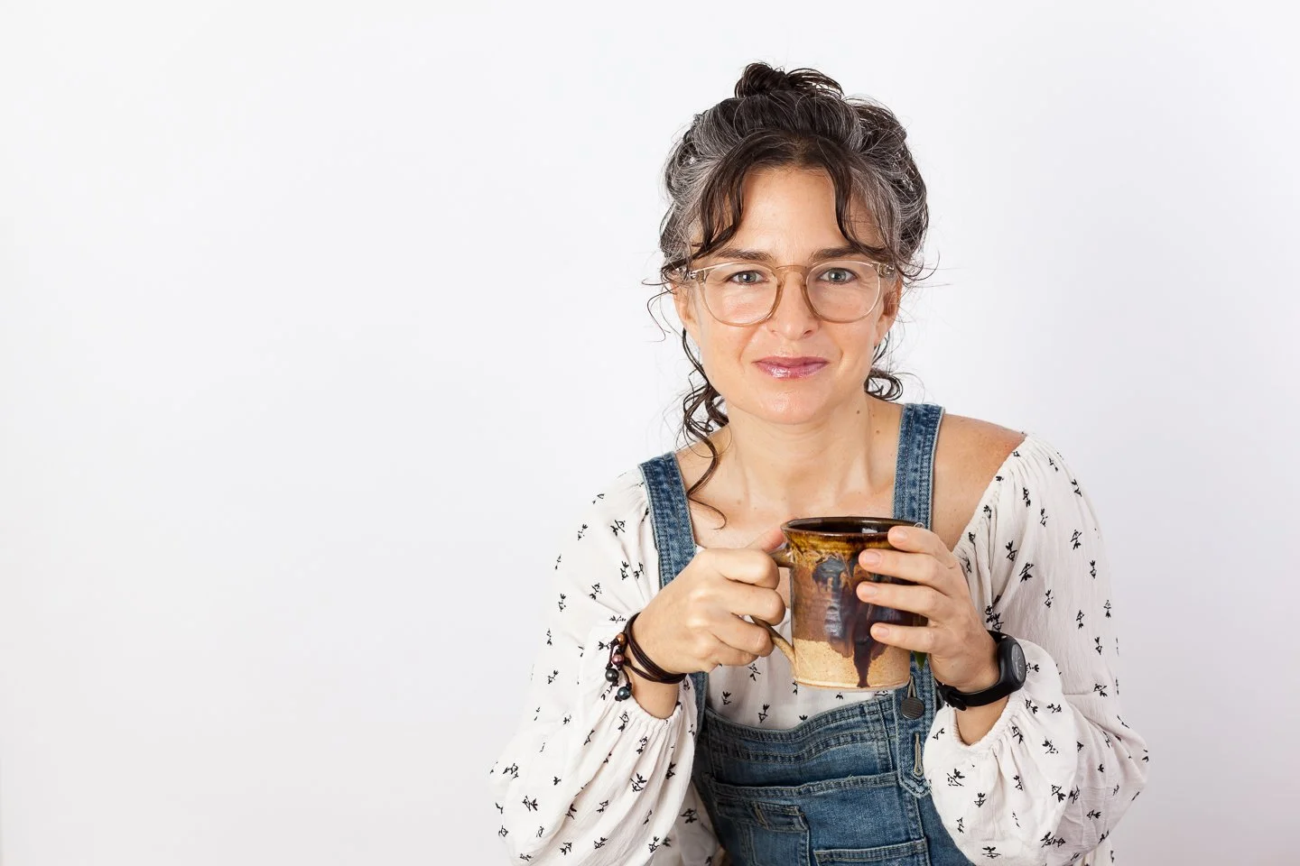 A woman with curly hair, glasses, and a watch, wearing a white blouse with small black patterns and denim overalls, holding a ceramic mug and smiling at the camera.