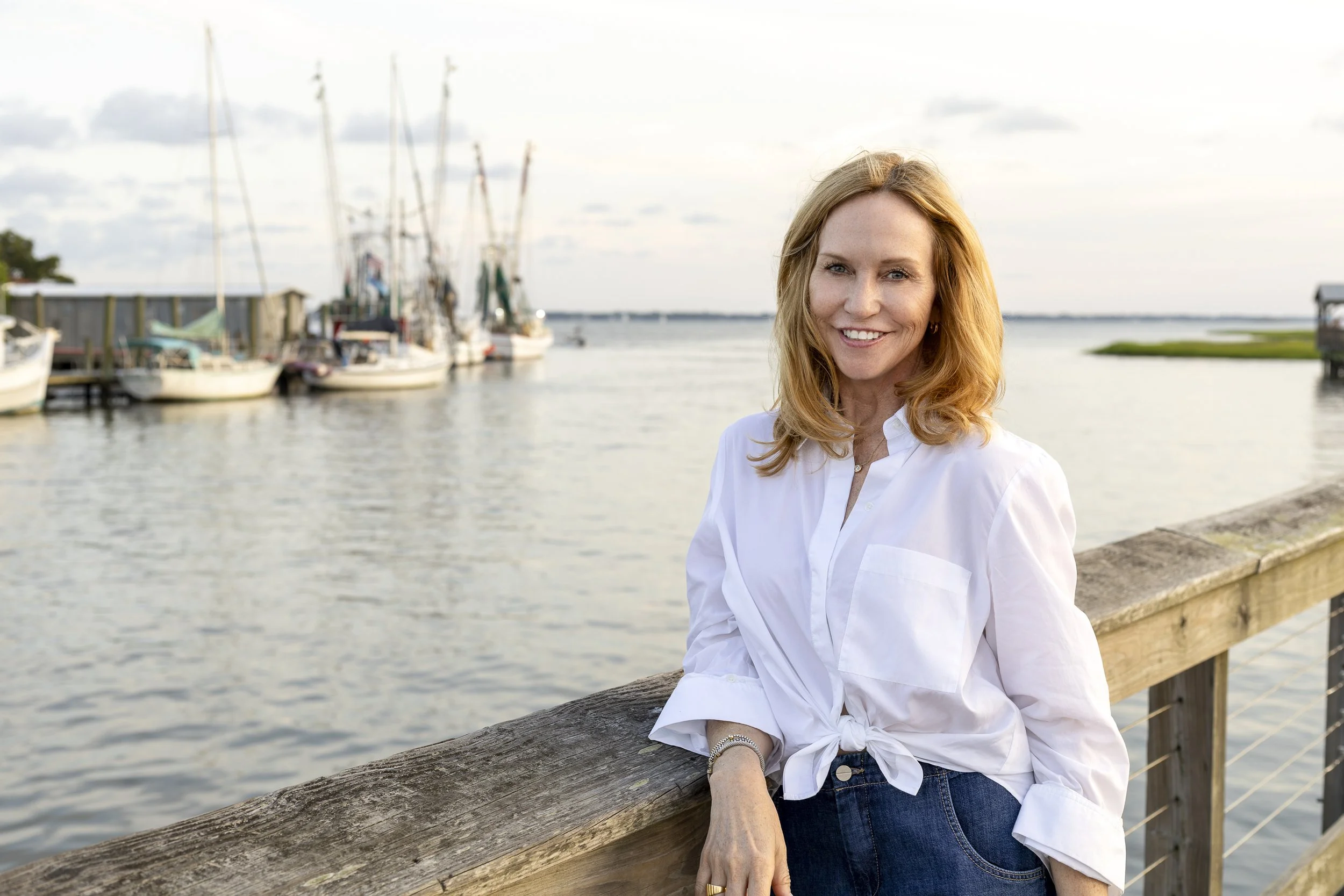 A woman with red hair and a white shirt standing on a wooden dock near a body of water, with sailboats and a cloudy sky in the background.
