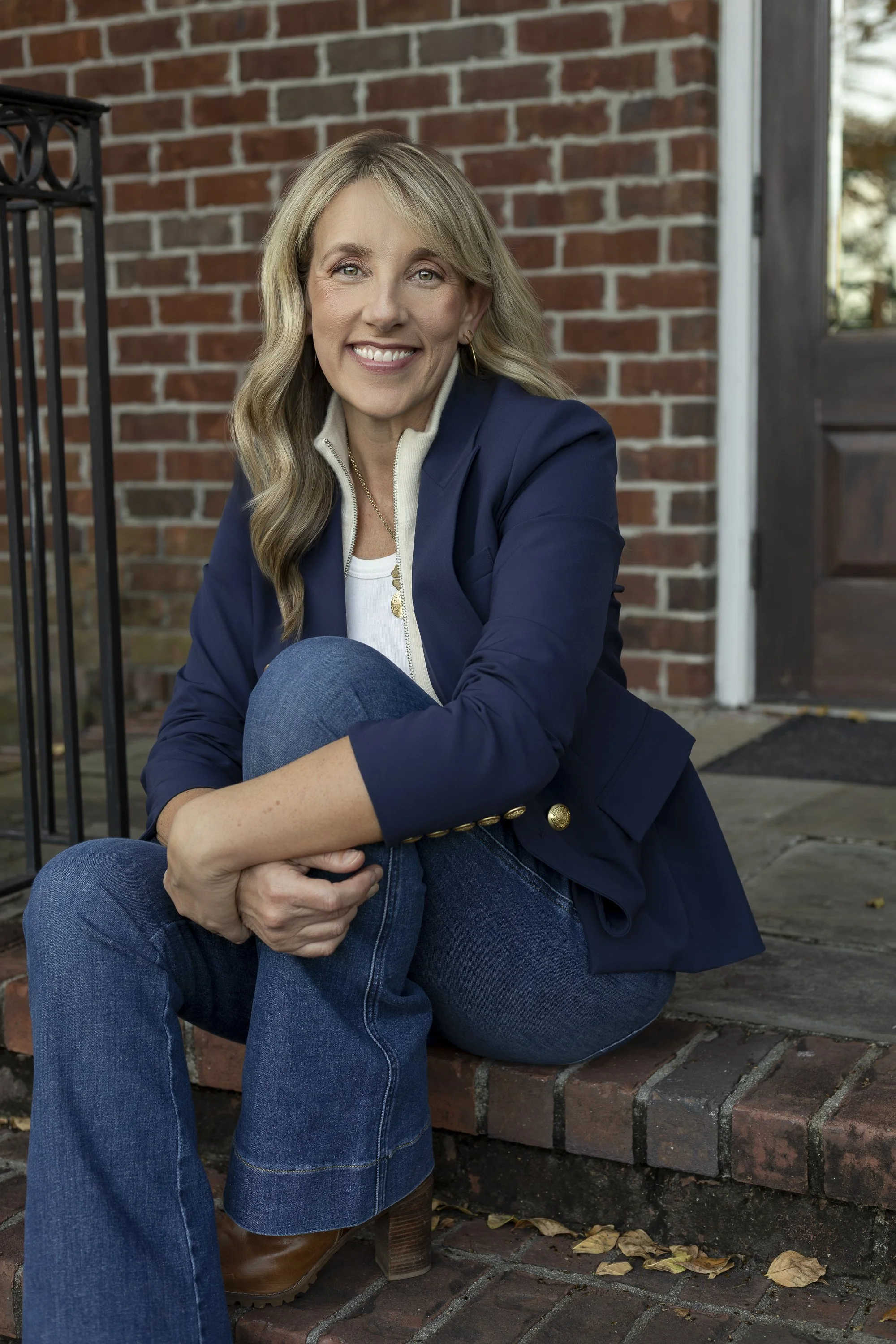 A woman with blonde hair sitting outside on a brick step, smiling at the camera, wearing a navy blazer, white top, and blue jeans.