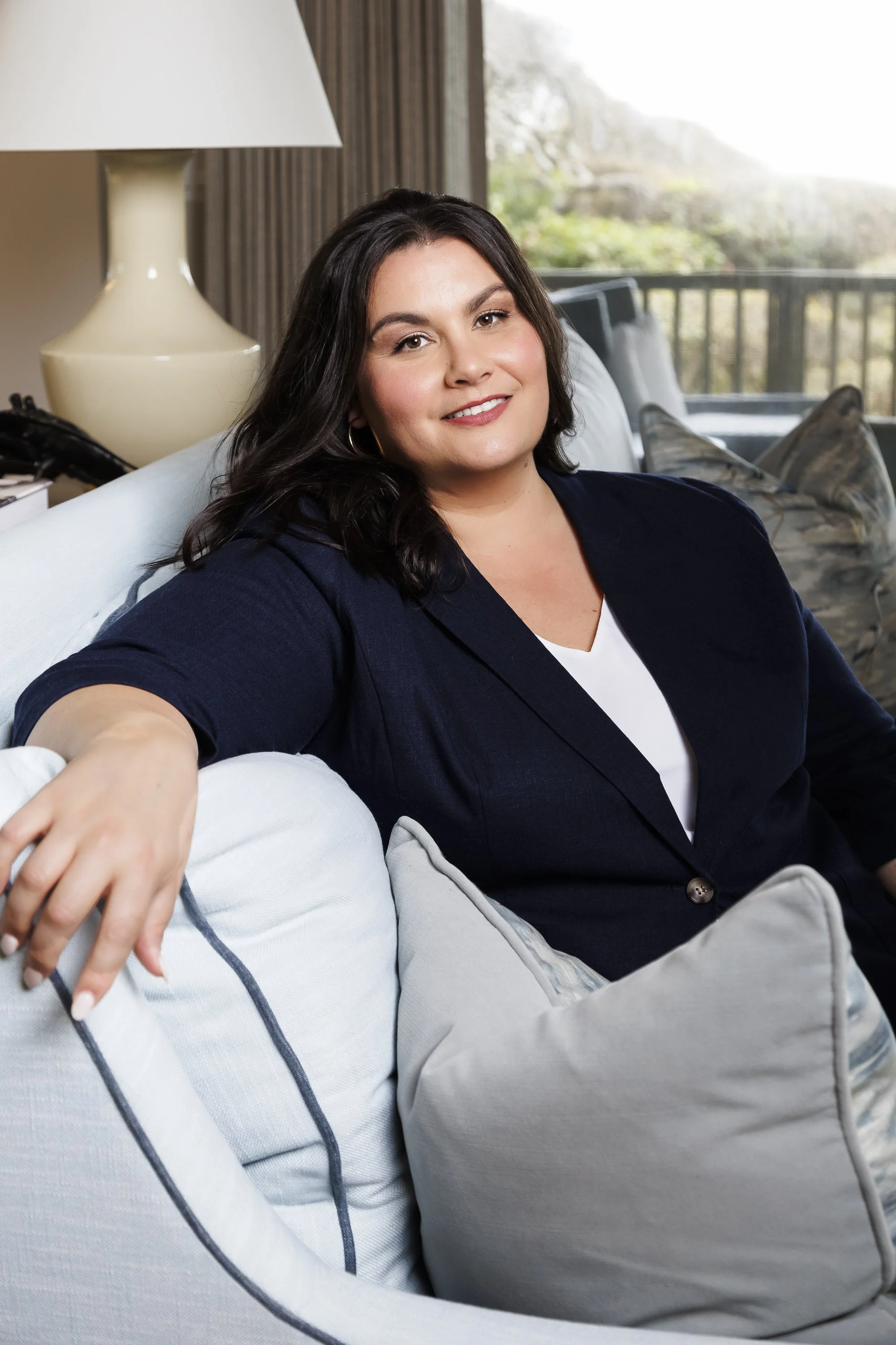 A woman with dark hair sitting on a light-colored couch in a well-lit room, smiling at the camera.