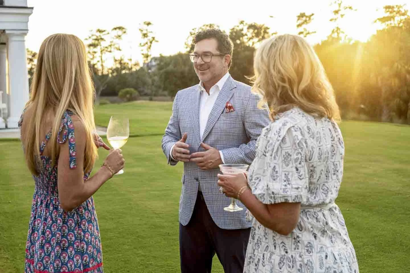 Three people having a conversation outdoors at sunset, holding wine glasses, in a garden area with a house in the background.