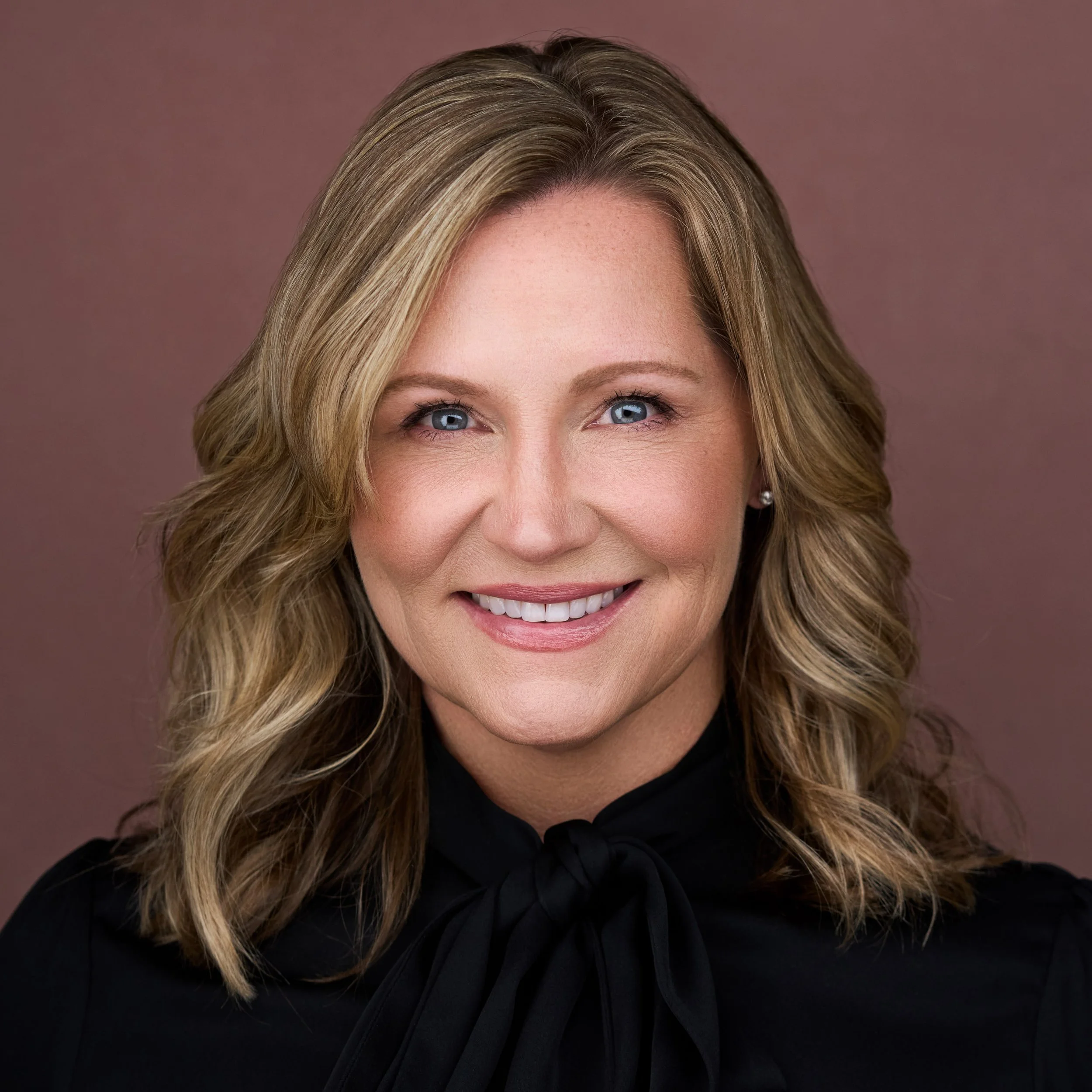 Close-up of a woman with blonde hair and blue eyes, smiling, wearing a black top with a high neckline, against a plain brown background.