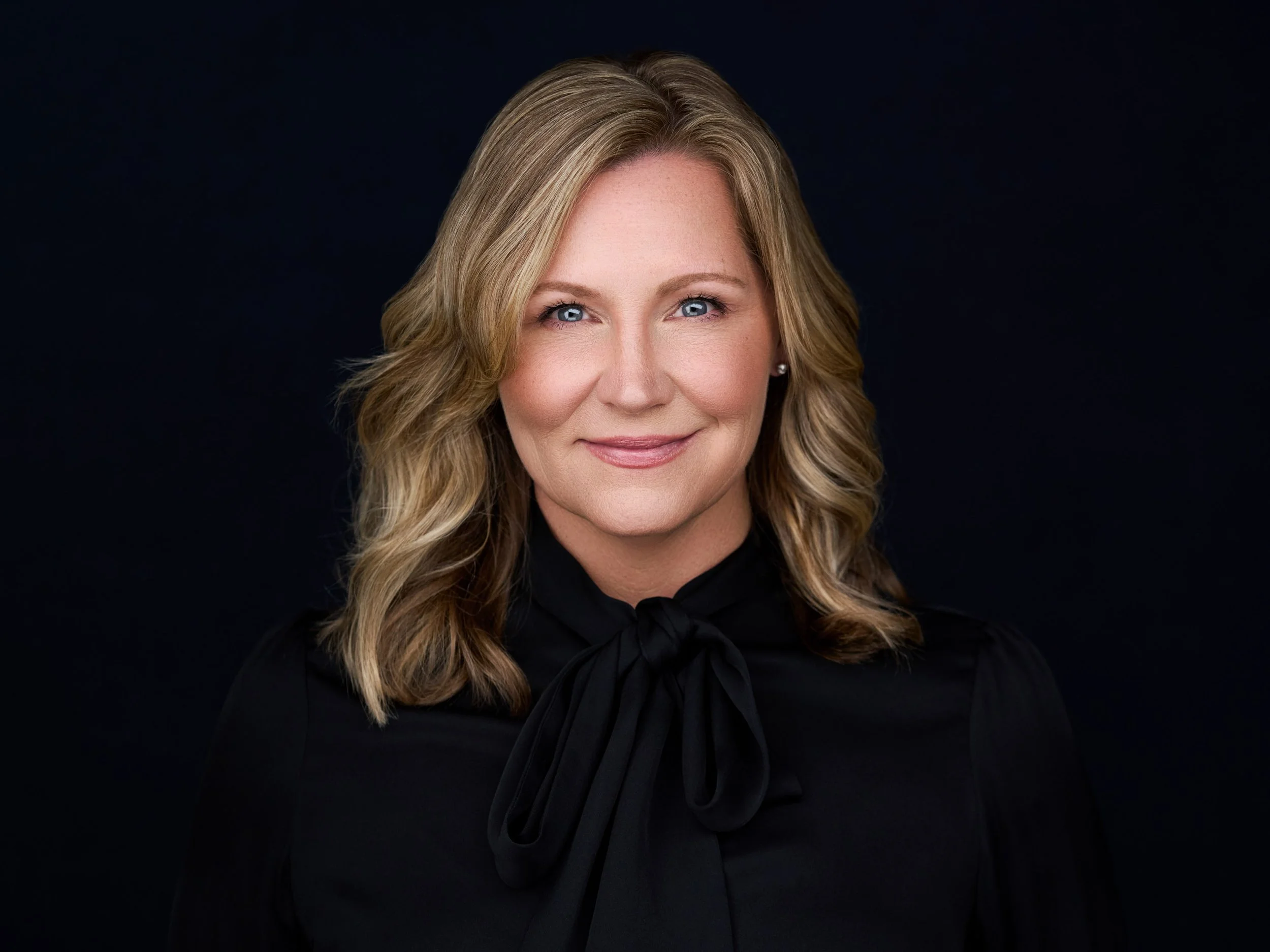 A woman with blonde wavy hair, blue eyes, and a slight smile, wearing a black top with a tied bow at the neck, against a dark background.