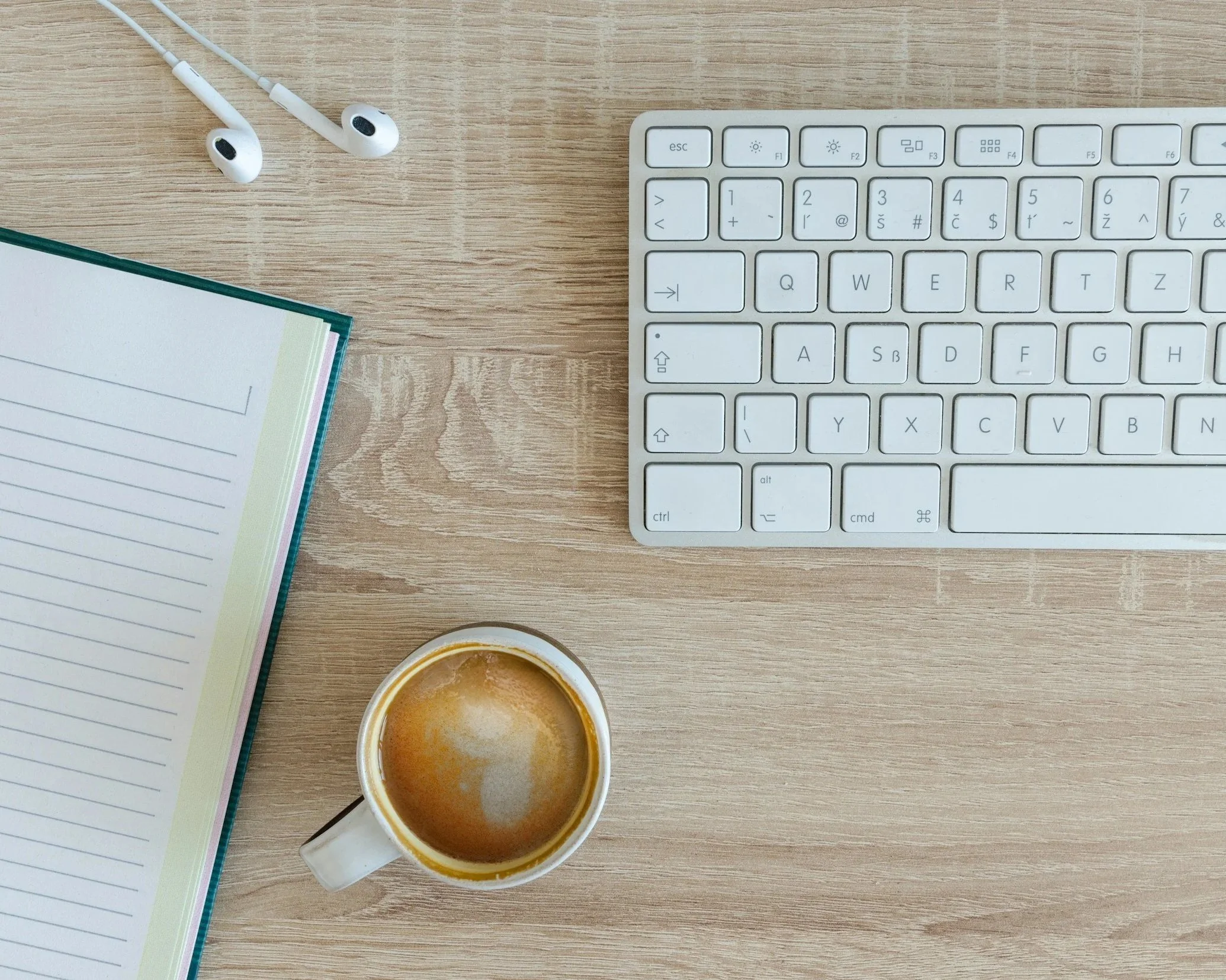A wooden desk with a white computer keyboard, a pair of white earphones, a notebook with lined pages, and a cup of coffee.
