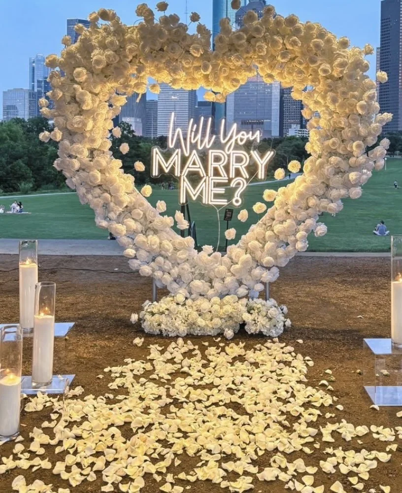 A large heart-shaped floral arrangement made of white roses, with a neon sign inside that reads, 'Will You Marry Me?'. Petals are scattered on the ground, flanked by candles on the sides, set outdoors with a city skyline and park in the background.