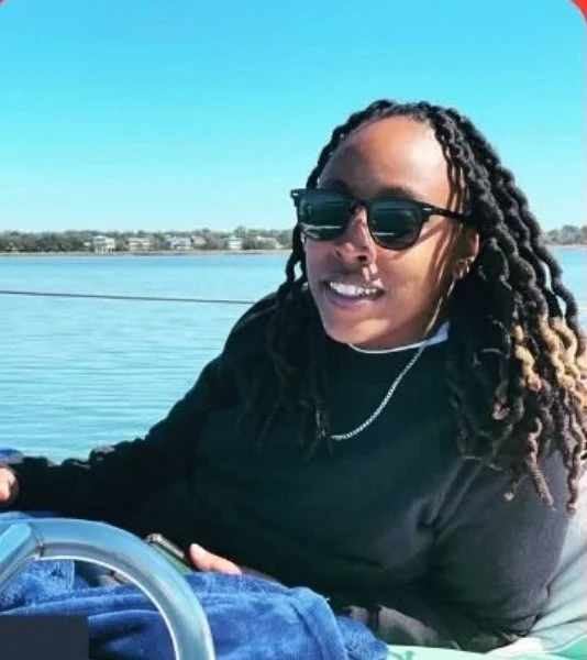 Woman with long dreadlocks, wearing sunglasses, smiling on a boat with water and shoreline in the background.