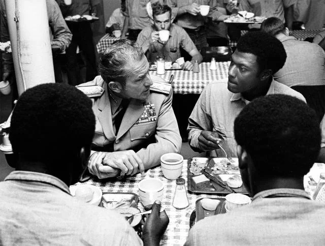 A black and white photo of a military officer having a serious conversation with a young man at a busy restaurant table, with other patrons in the background.