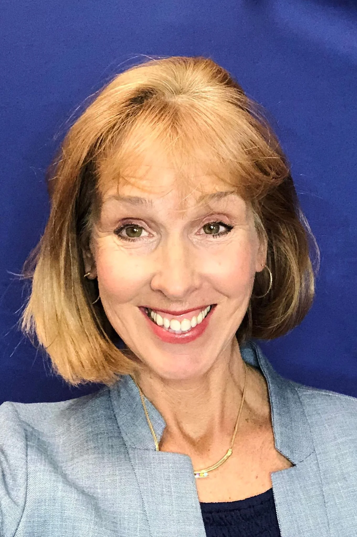A smiling woman with shoulder-length red hair, wearing a gray jacket, a black top, and gold jewelry, standing in front of a blue background.