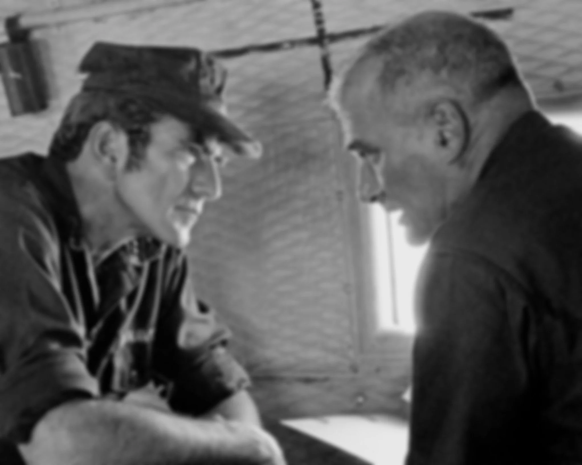 Two men are sitting and leaning towards each other, smiling and appearing to share a moment of laughter. They are indoors, near a window with blinds.