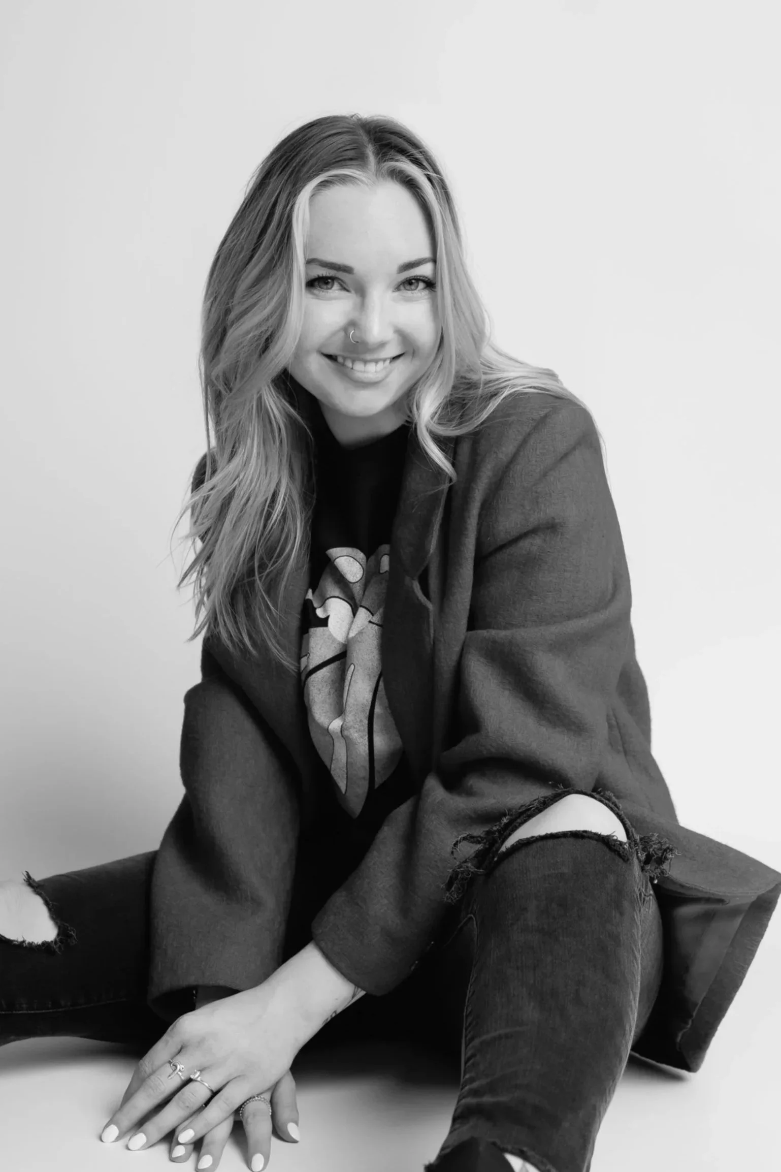 Black-and-white photo of a young woman sitting on the floor, smiling at the camera with her legs apart and one hand resting on her knee.