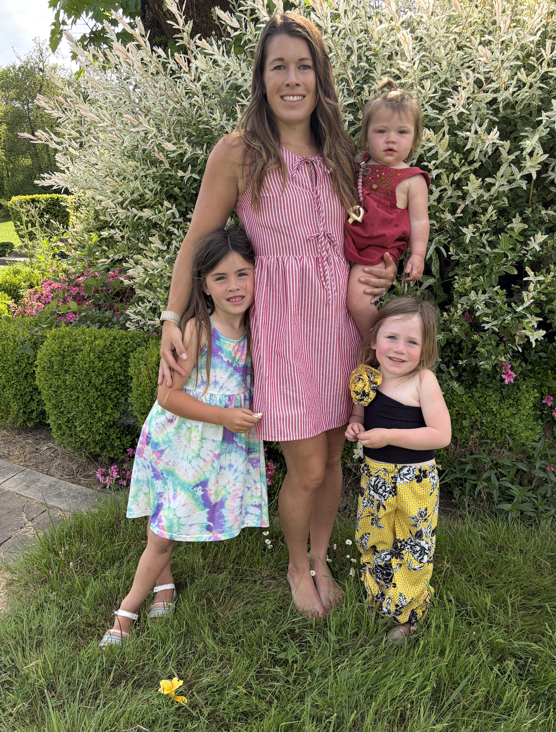 A woman with four young girls stands outdoors in front of green bushes and flowering plants, smiling at the camera.