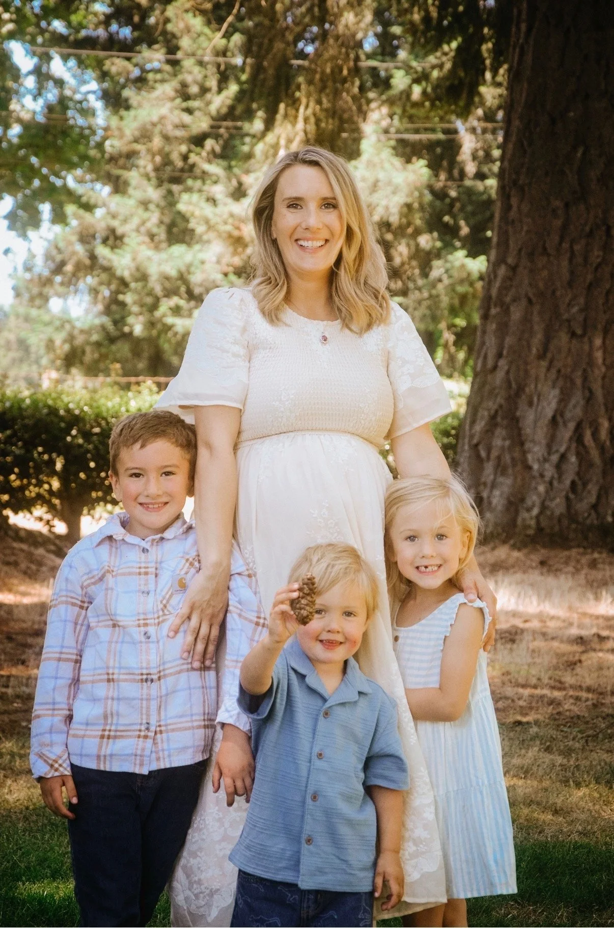 A woman in a white dress standing outdoors with four children in front of her, near a large tree and greenery.