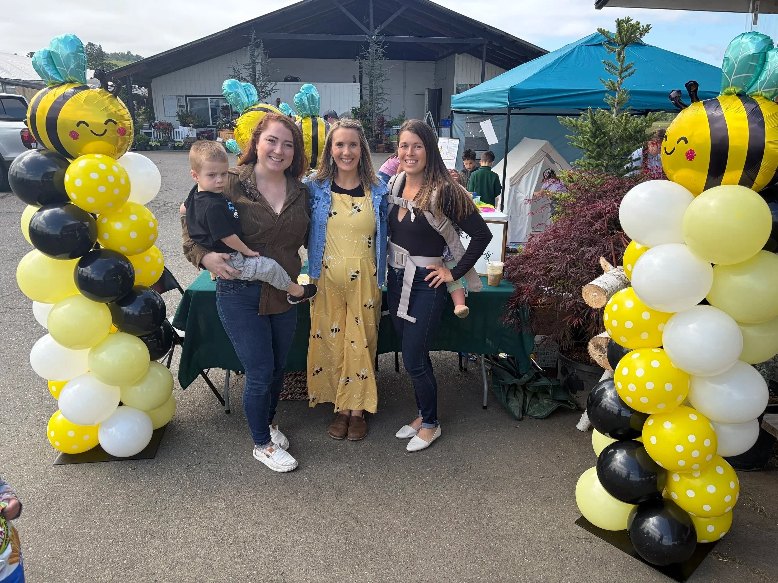 Three women and a child standing together under a balloon arch with smiling bee faces, at an outdoor event with tents and people in the background.