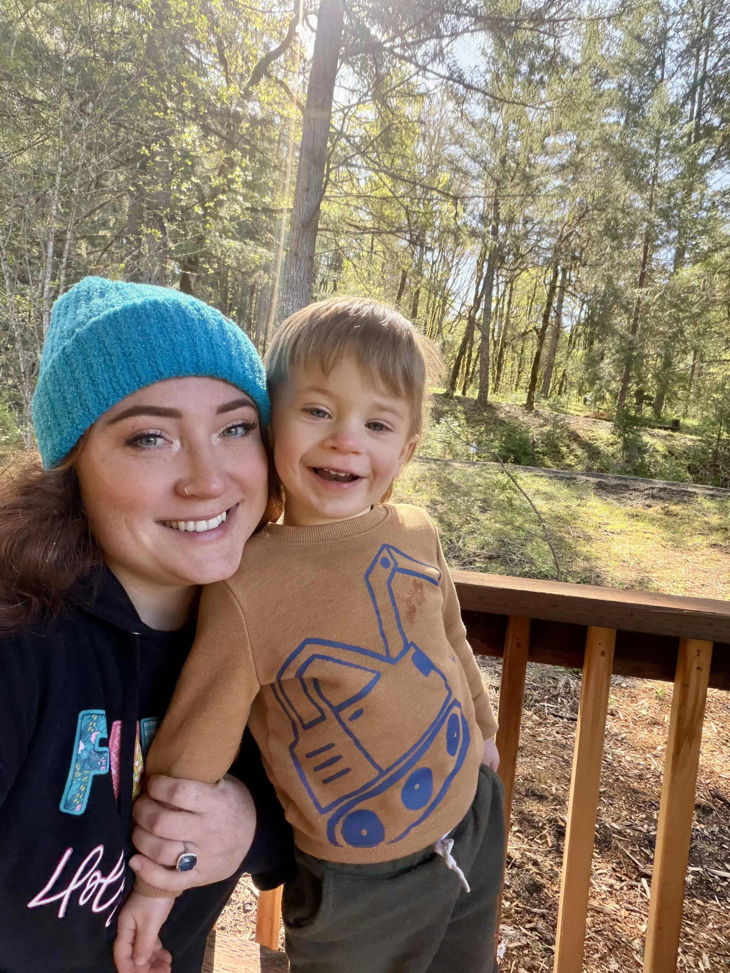 A woman with long red hair, wearing a blue knit hat, and a young boy with short light brown hair, smiling, standing outdoors on a wooden deck with a forest background of tall trees and sunlight filtering through the leaves.