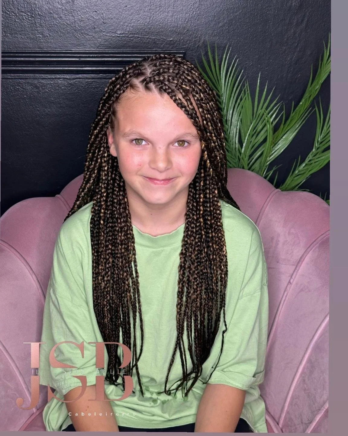 A young girl with long, brown braids, wearing a light green t-shirt, sitting on a pink chair in front of a dark wall with a large green plant behind her.