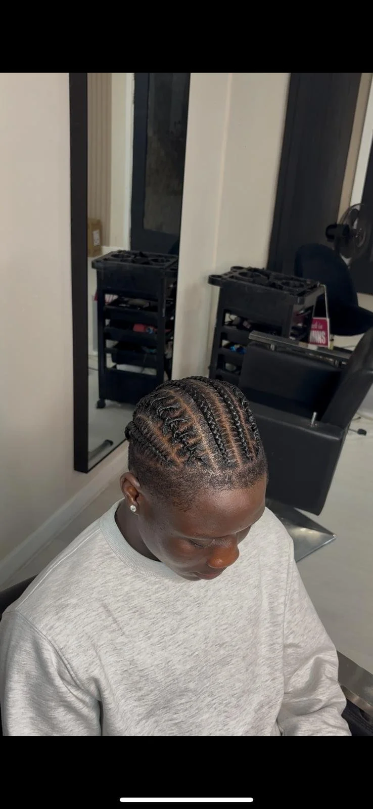 A woman with braided hairstyle sitting in a hair salon, with mirrors and hair styling tools visible in the background.