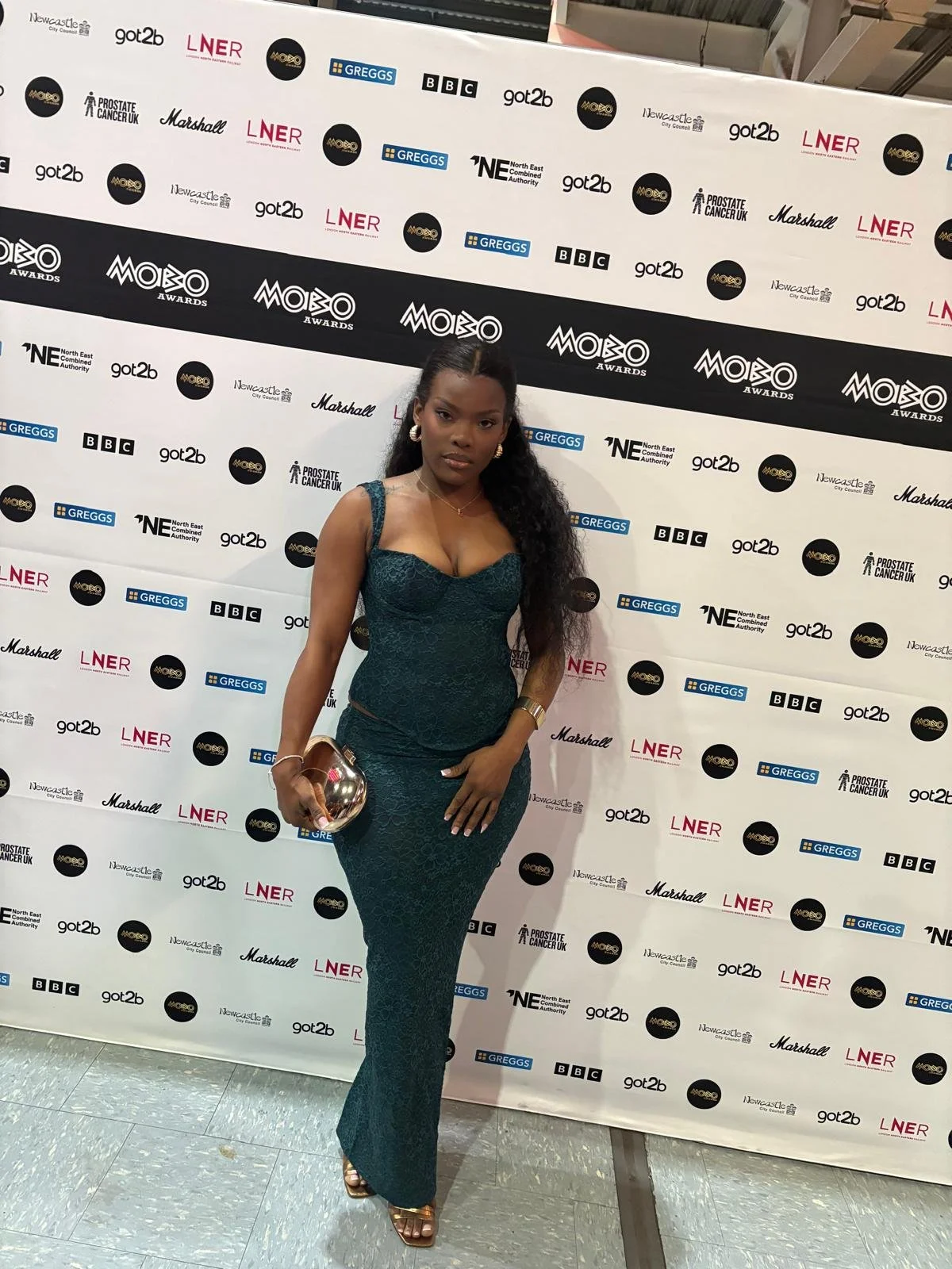 A woman in a teal lace dress standing on a red carpet in front of a step and repeat banner at an awards event, holding a metallic clutch purse.