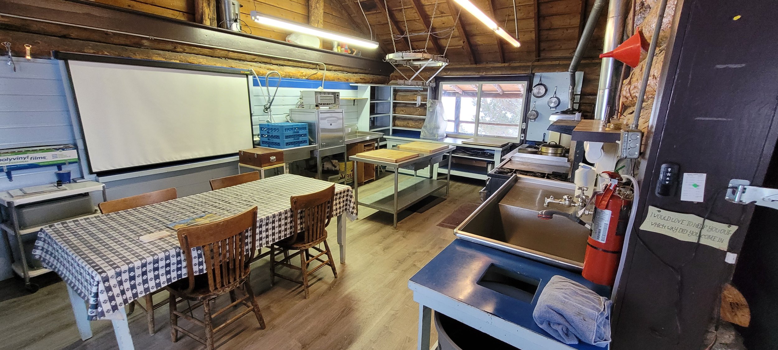 Interior of a rustic kitchen or workshop with a large white projection screen on the wall, a checkered tablecloth on a dining table, wooden chairs, metal work tables with cutting boards, pots and pans, a window letting in natural light, a fire extinguisher, cleaning supplies, and a fire hat on the wall.